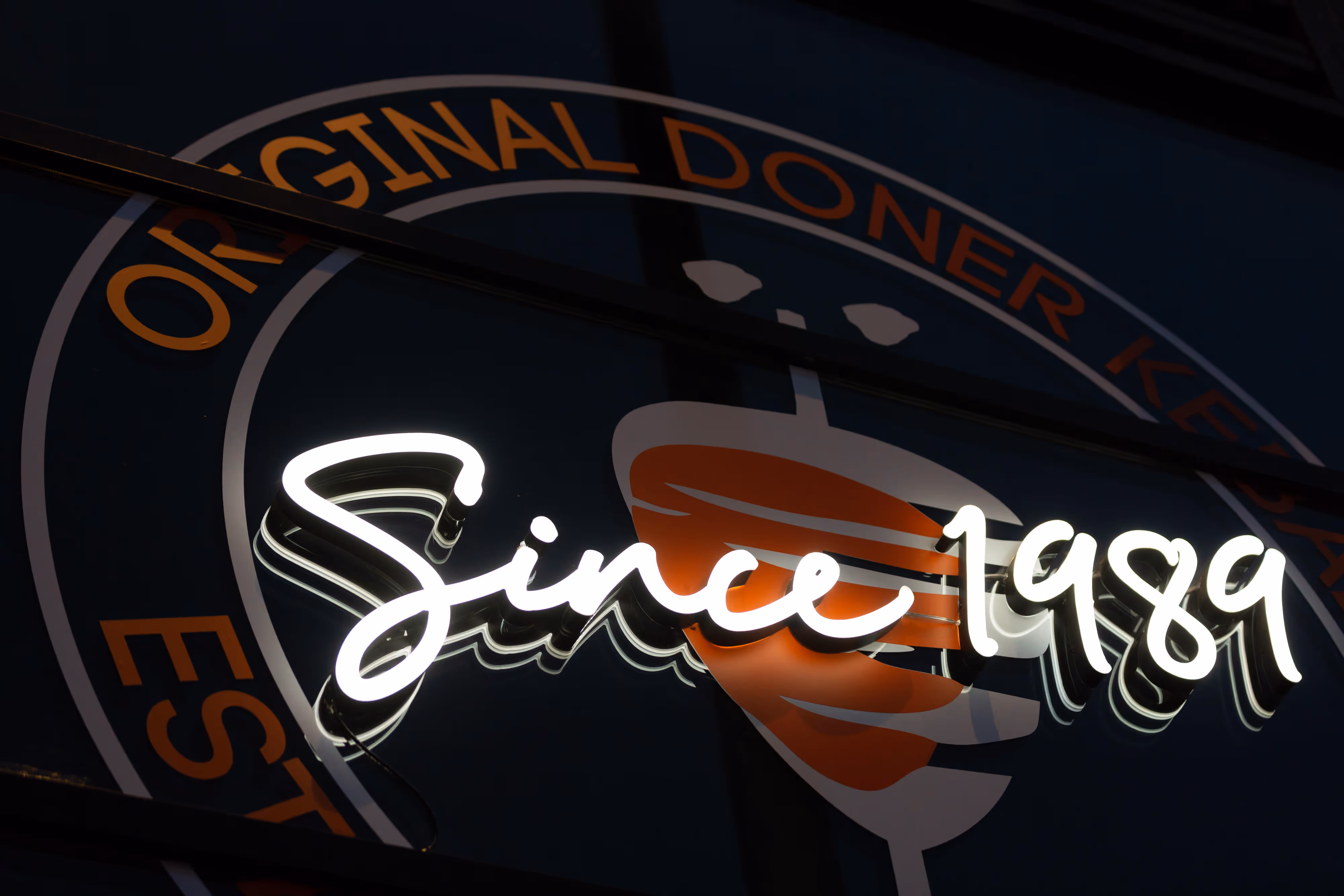Illuminated neon sign stating "Since 1989" positioned above the doner kebab restaurant’s original logo at shopfront exterior.