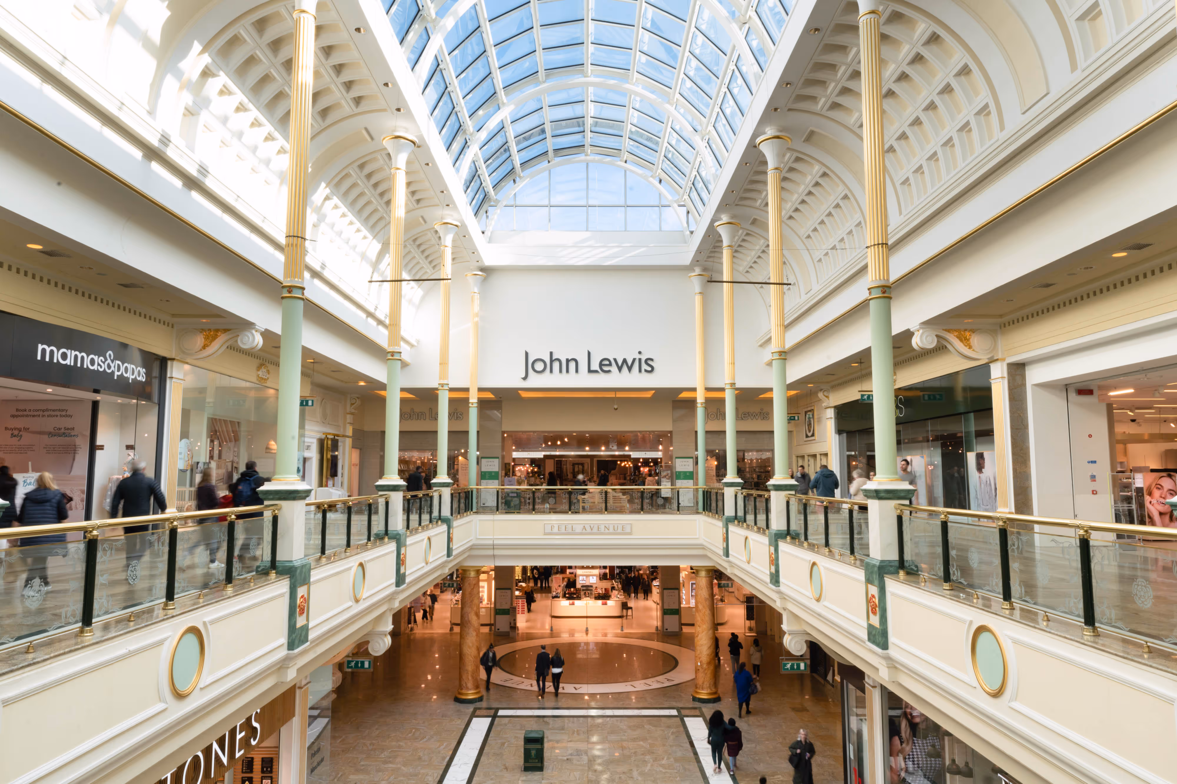 Wide shot of a vibrant shopping centre interior, glass ceiling overhead, with John Lewis shopfront and shoppers moving through the space.