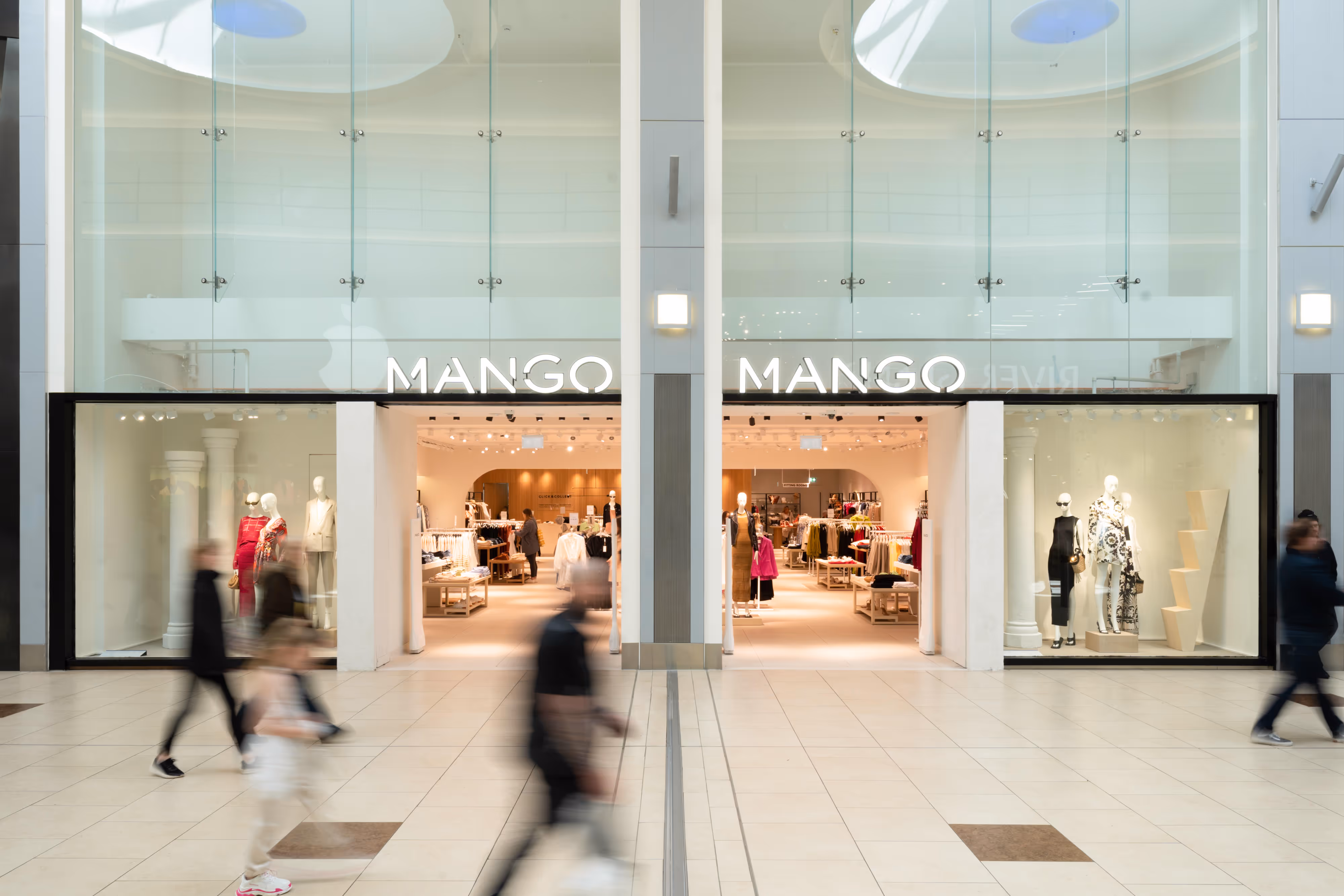 Photograph of Mango shop entrance in a shopping centre, featuring passing shoppers and mannequins displayed inside the well-lit window.