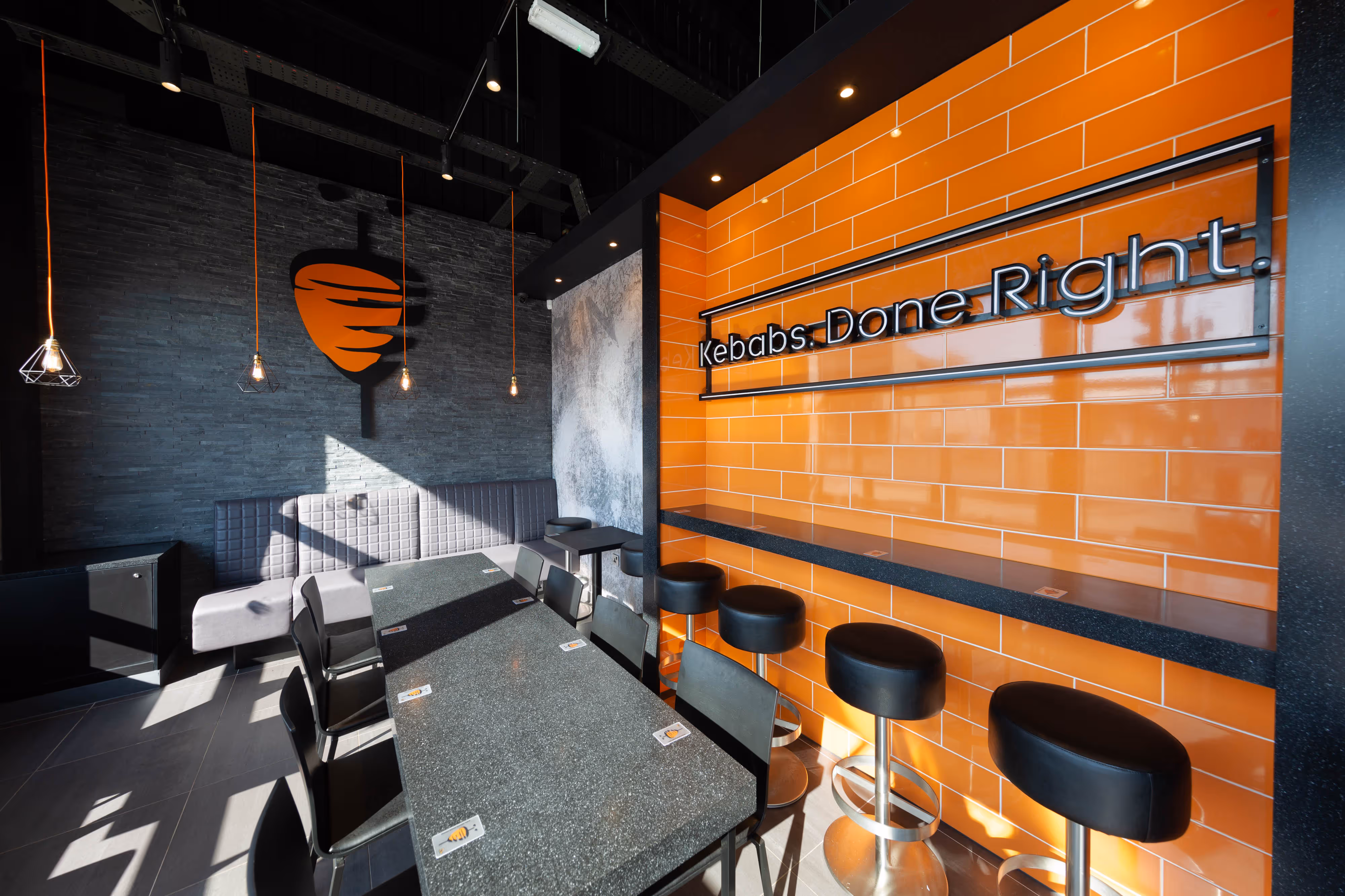 Capturing a vibrant kebab shop interior with bold orange tiles, sleek black stools, and a glowing “Kebabs. Done. Right.” neon sign.