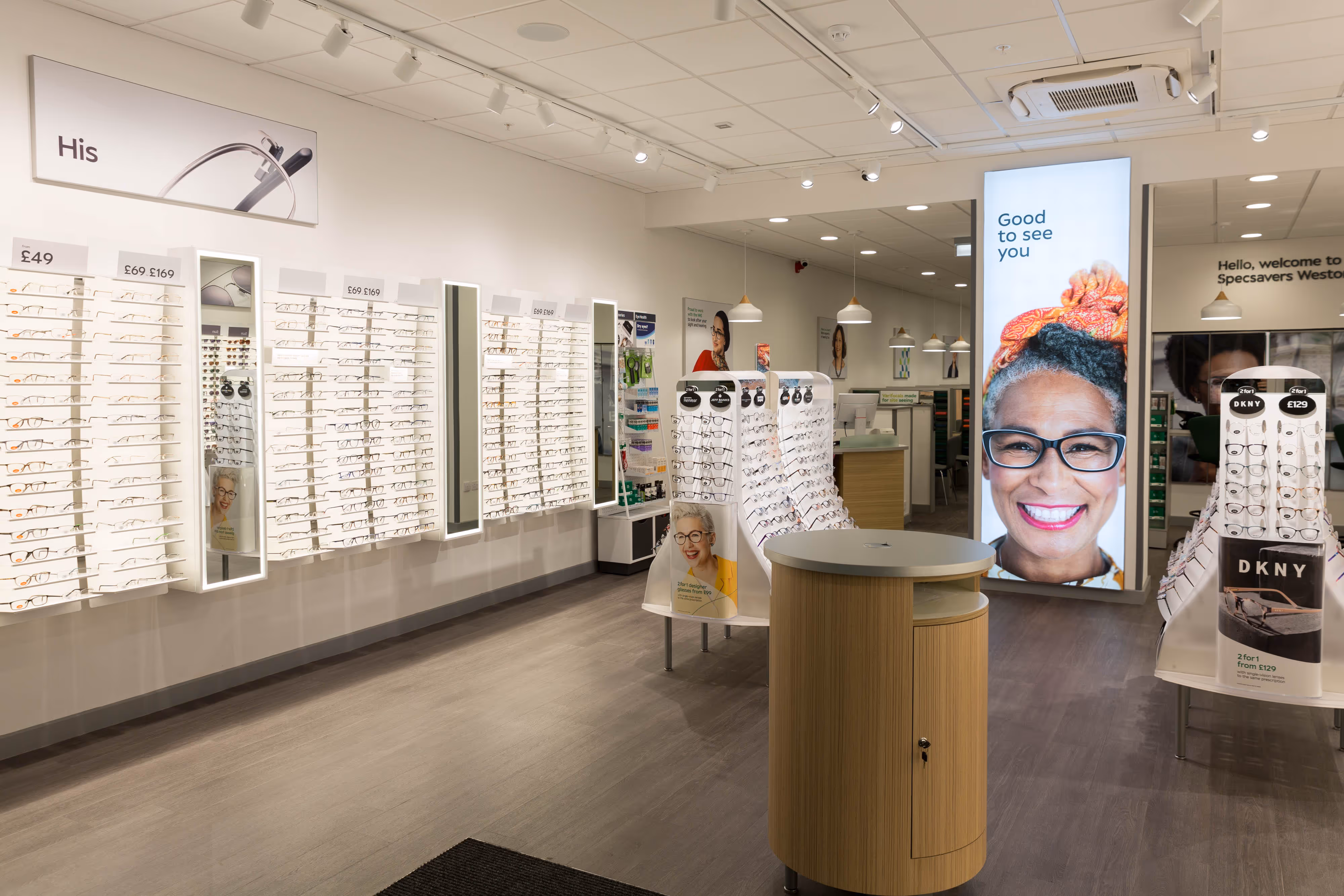 Spectacle frames arranged in a sleek optician's shop, captured with a prominent "Good to see you" sign and smiling woman.