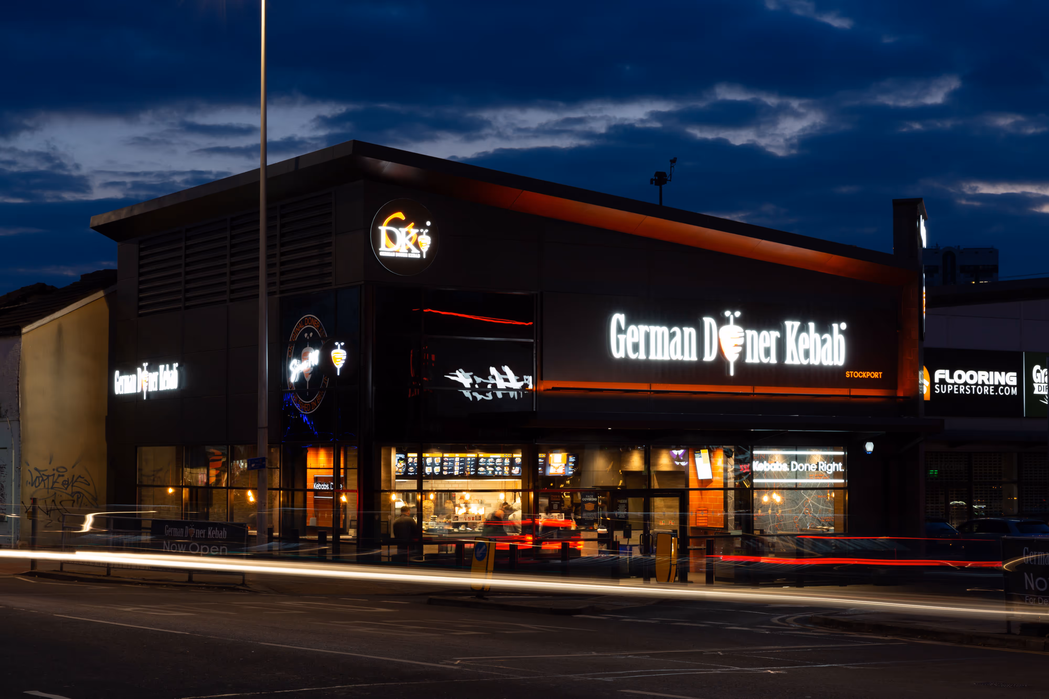 German Doner Kebab shopfront illuminated at night, lively ambience highlighted by passing car lights on the busy street outside.