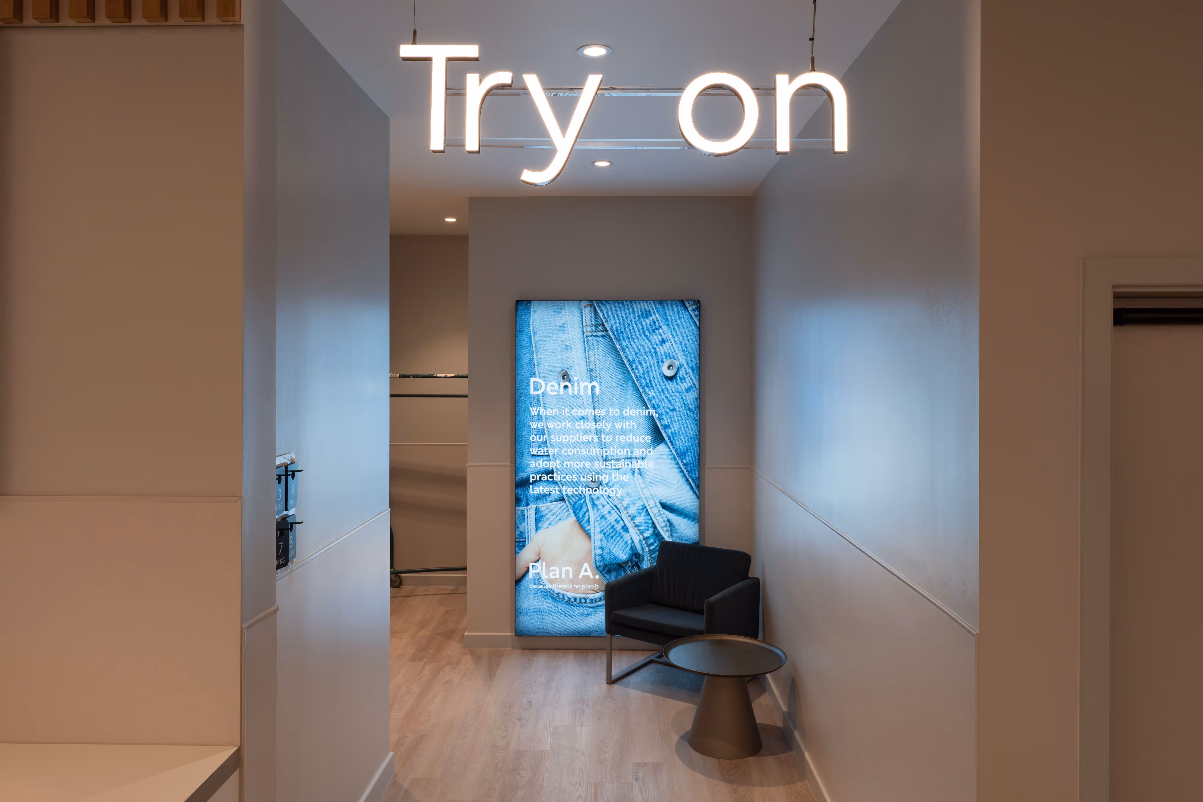 Fitting room setup: neon “Try on” sign glows above denim-themed display, chair, and table - captured in an M&S retail outlet..