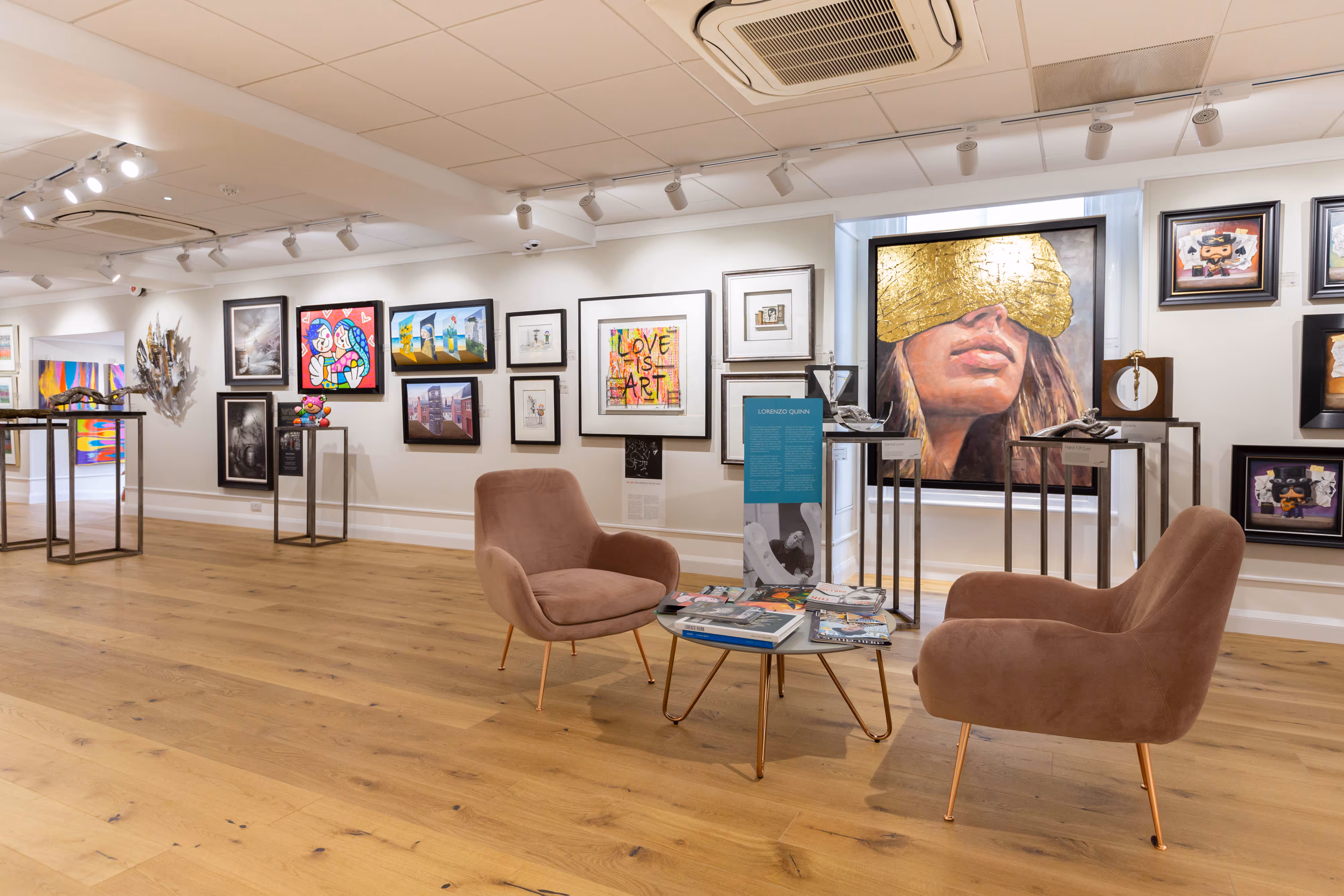 Retail-style display with framed modern art on white walls, two brown chairs, a glass table, and polished wooden floors.