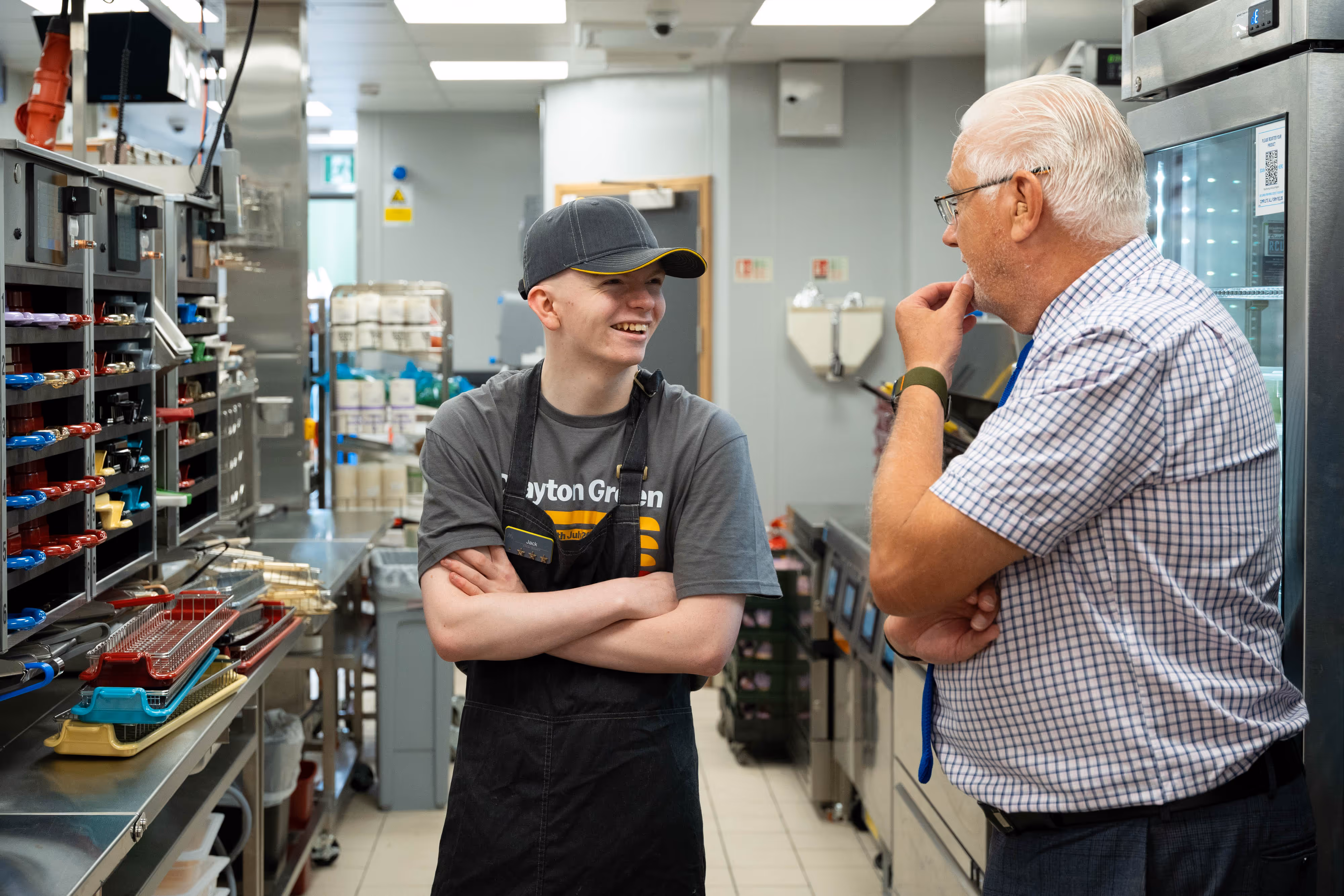 Young employee in a McDonald's uniform chats and smiles with older manager in shirt and tie in a commercial kitchen.