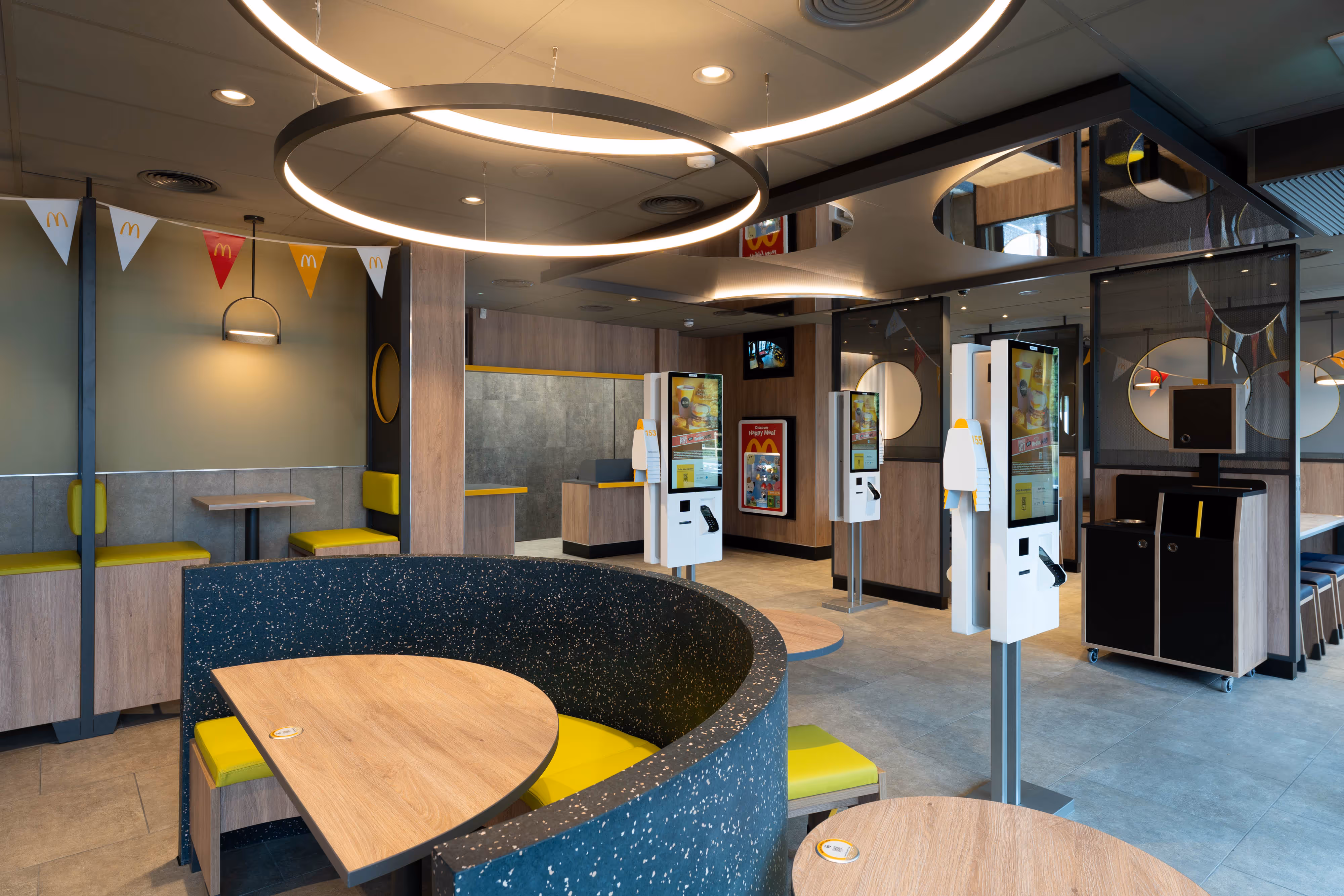 Photograph of a McDonald's interior featuring wooden tables, yellow seating, digital ordering kiosks, and stylish lighting.