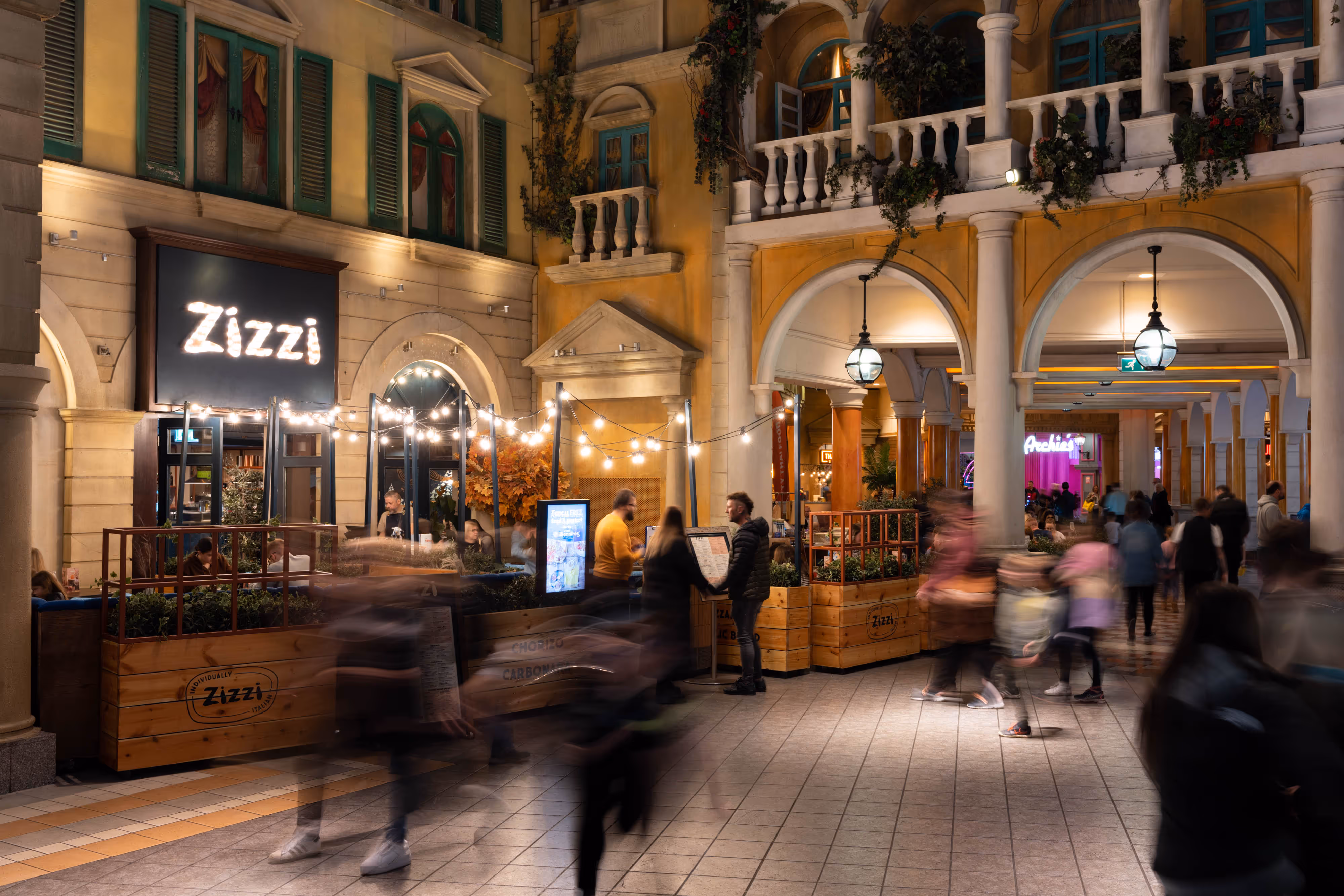 Shoppers stroll by a restaurant with fairy-lit outdoor seating, set in a vibrant, European-inspired indoor retail arcade.