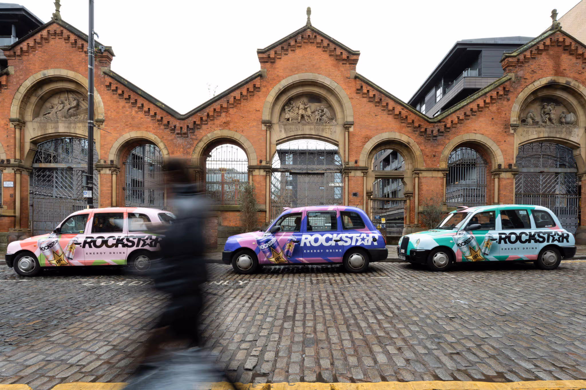 Framed three vibrant taxis, each wrapped in bold "Rockstar" adverts, lined up on cobblestones as a passer-by animates the scene.