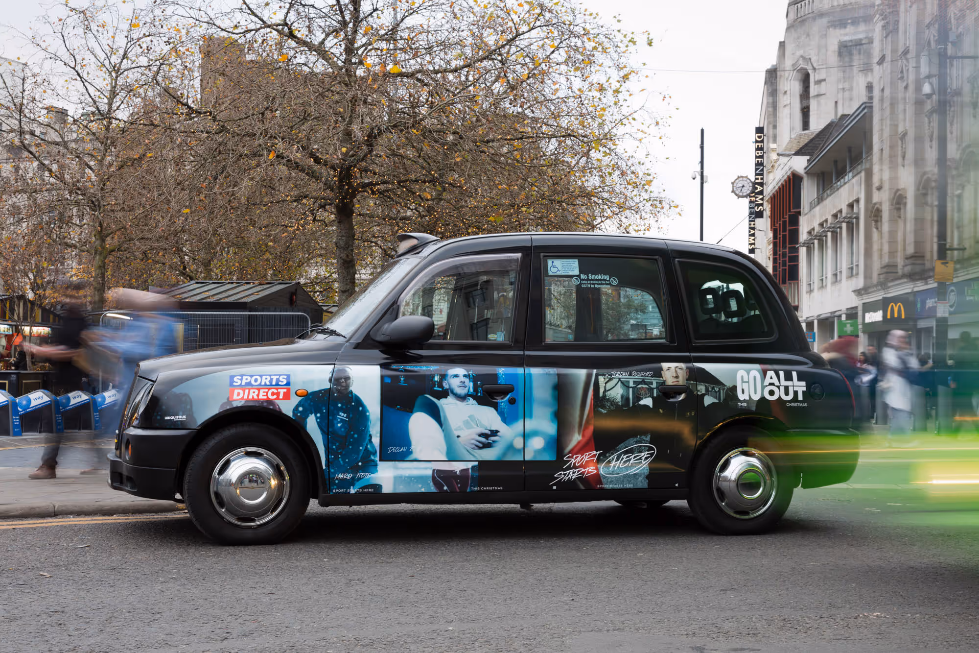Captured a black cab wrapped in bold sports-themed adverts, stationed on Oldham Street with bare trees and urban architecture.