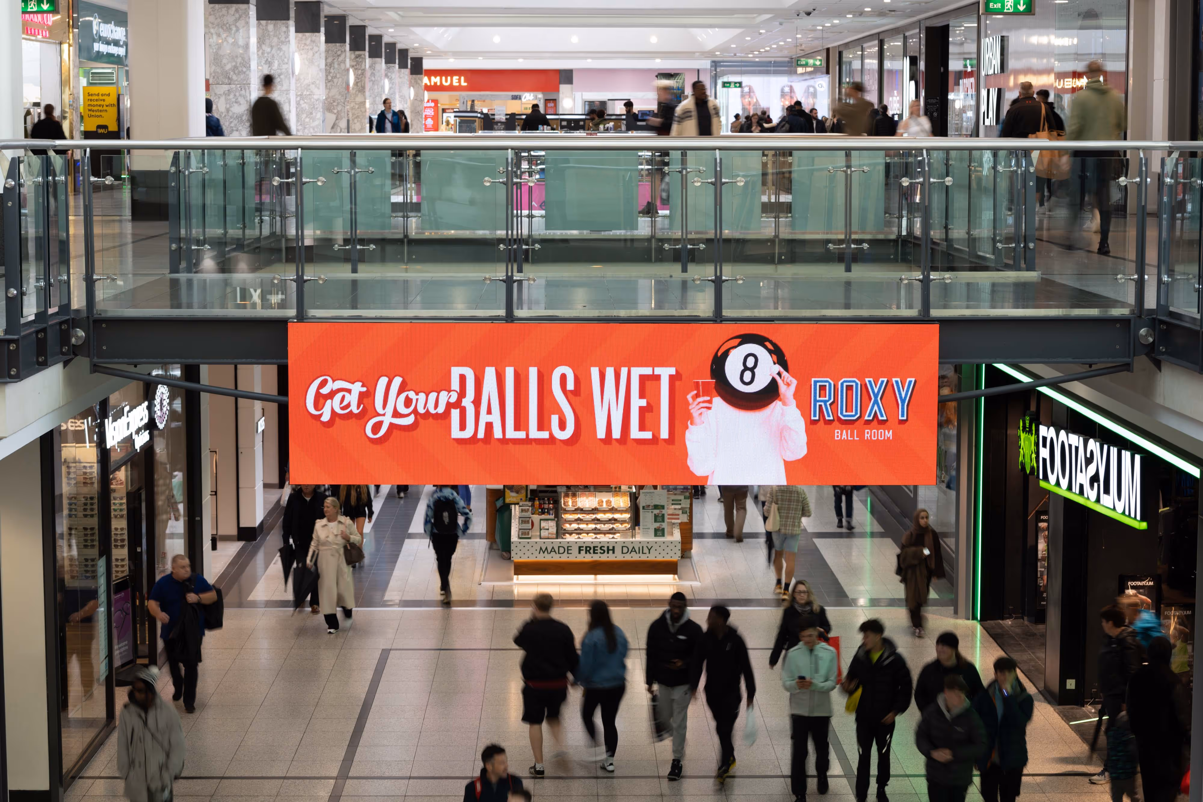 Documenting the Arndale Centre floor, I capture shoppers moving beneath a bold red "Get Your Balls Wet at Roxy" banner overhead.