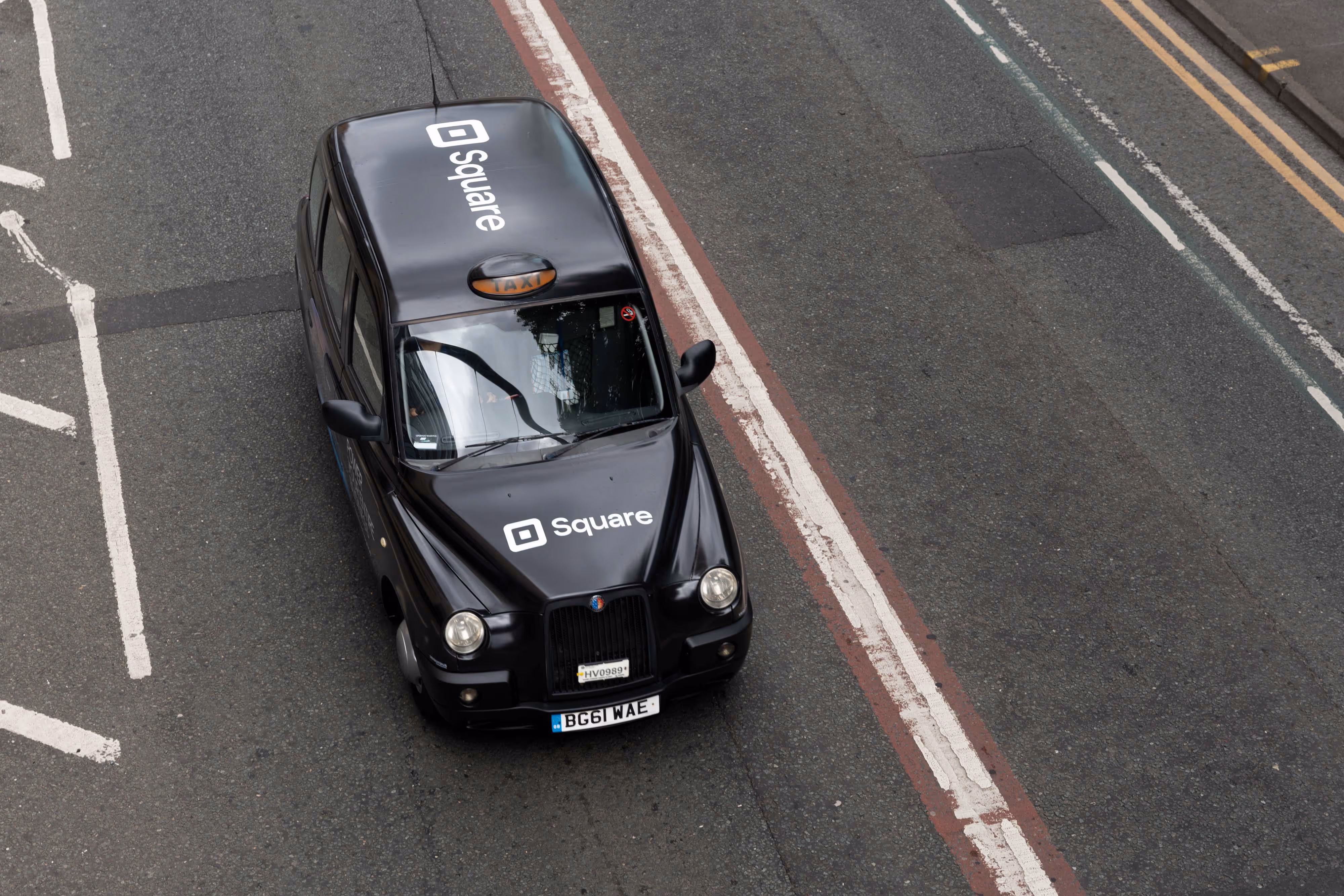 Captured through my lens, a black taxi adorned with Square branding cruises past bold red and white lane markings in the city centre.