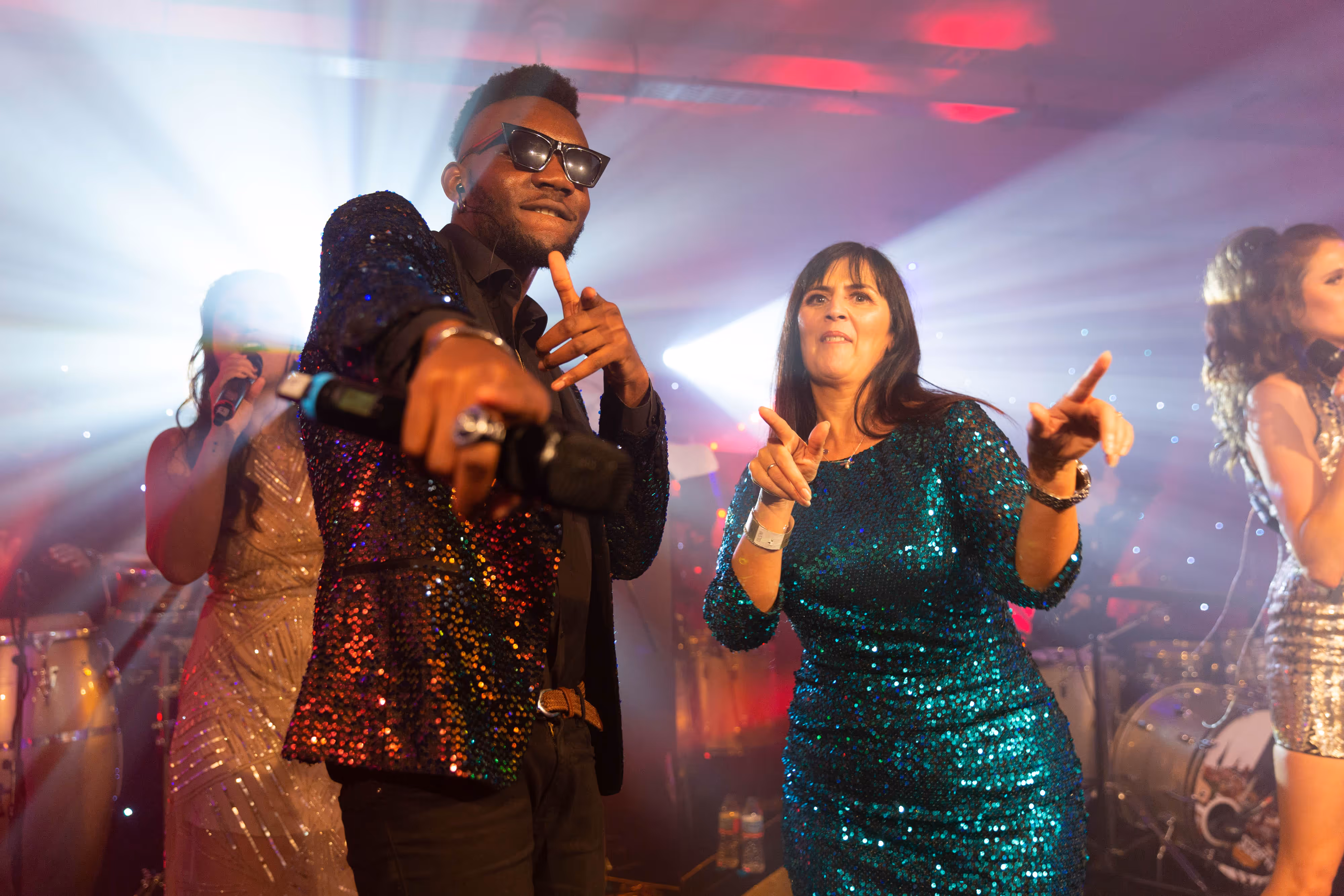 Team members in glittering outfits dance and point at the camera on stage under bright lights at a Manchester Christmas do.