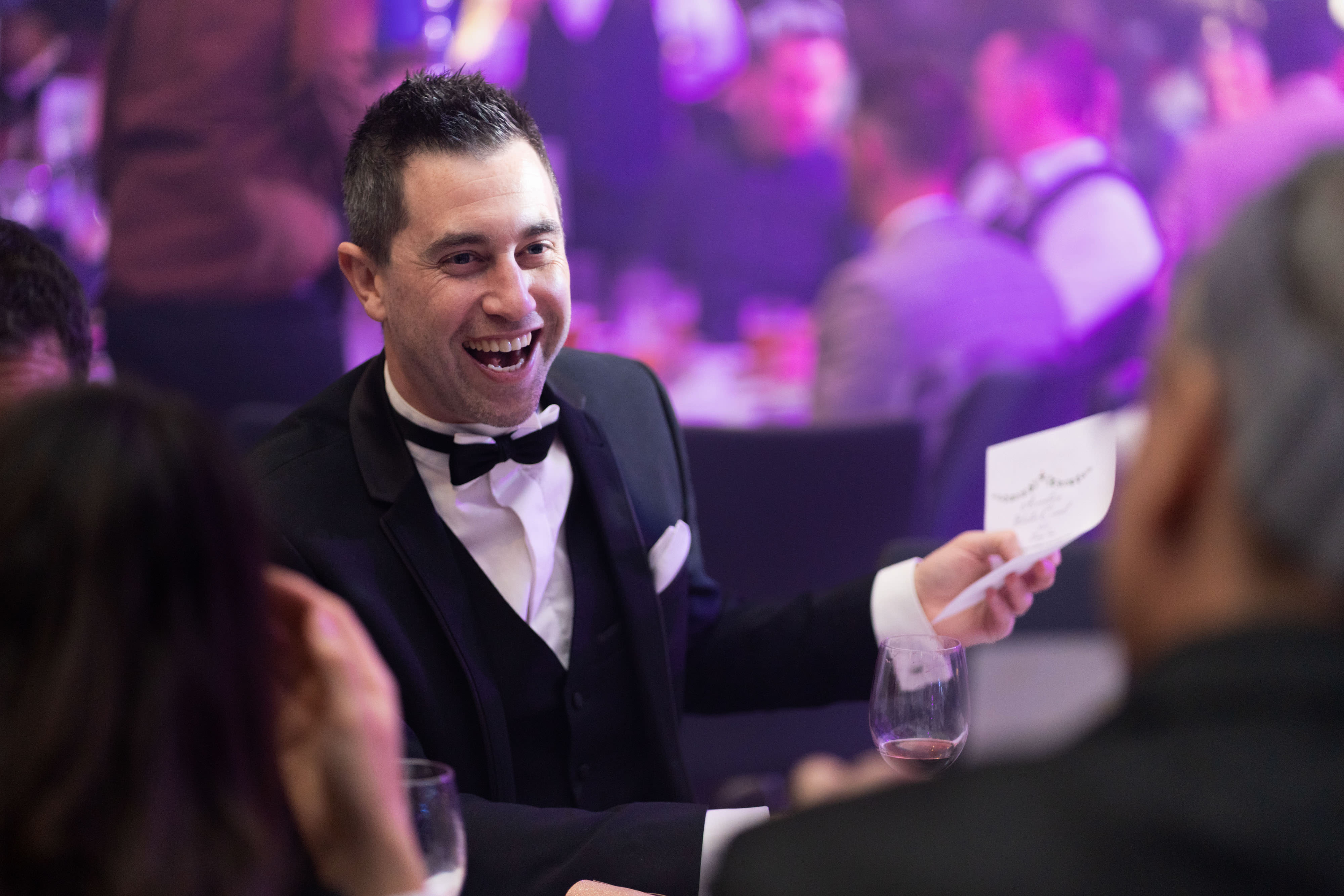 A guest in a dinner jacket holding a card and wine glass among festive colleagues.
