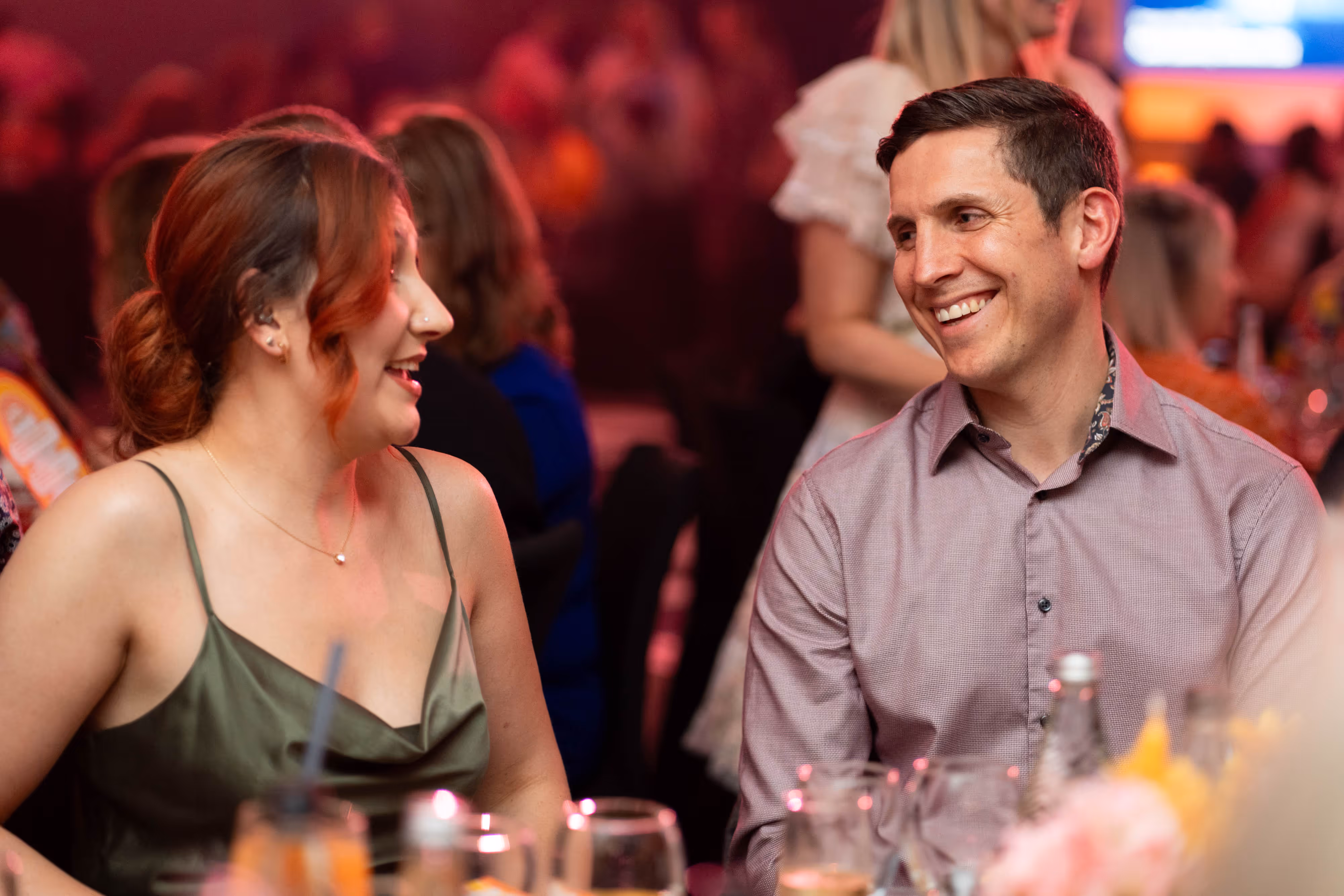 A man and woman share a laugh at their table, with the lively crowd behind them.