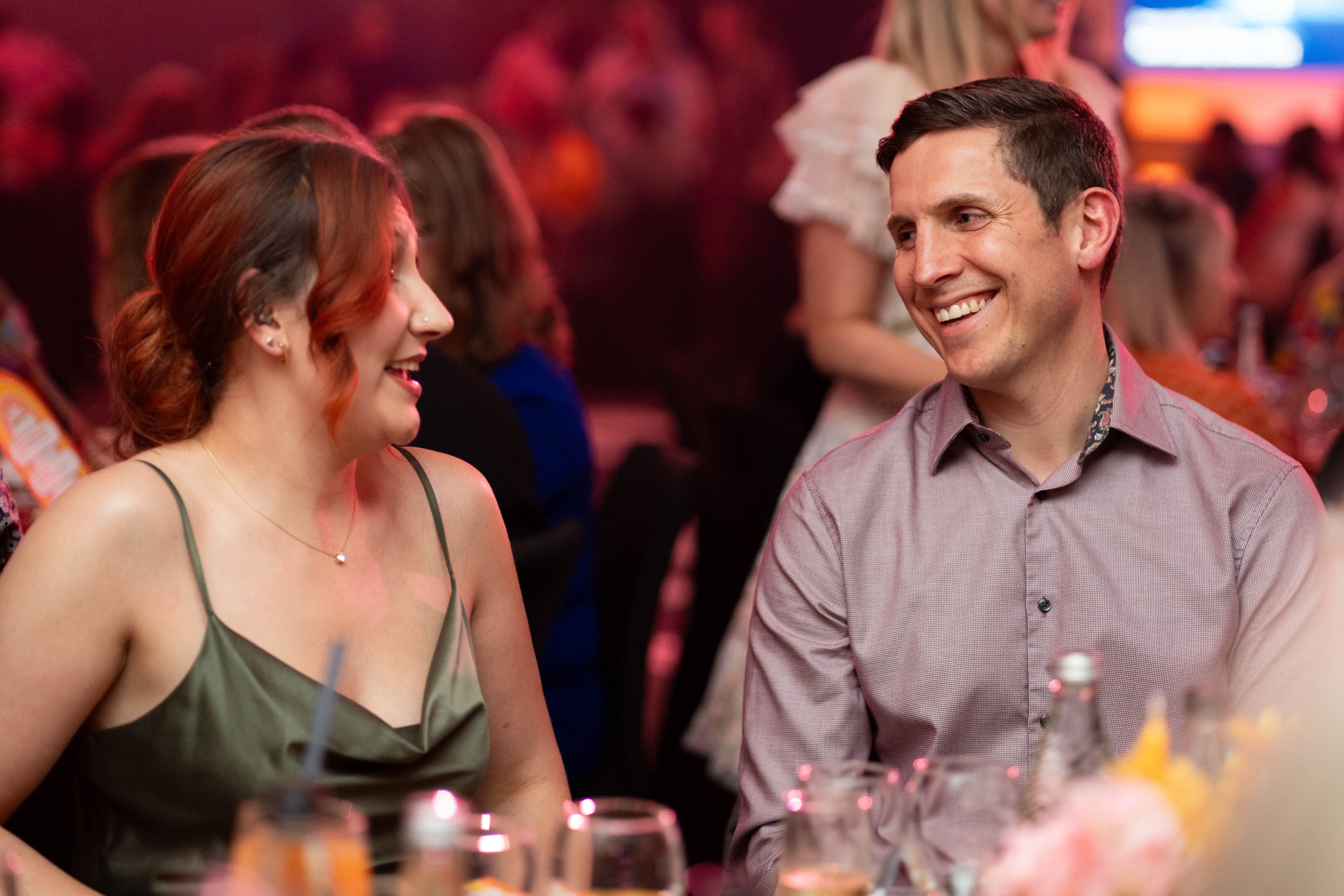 A man and woman share a laugh at their table, with the lively crowd behind them.