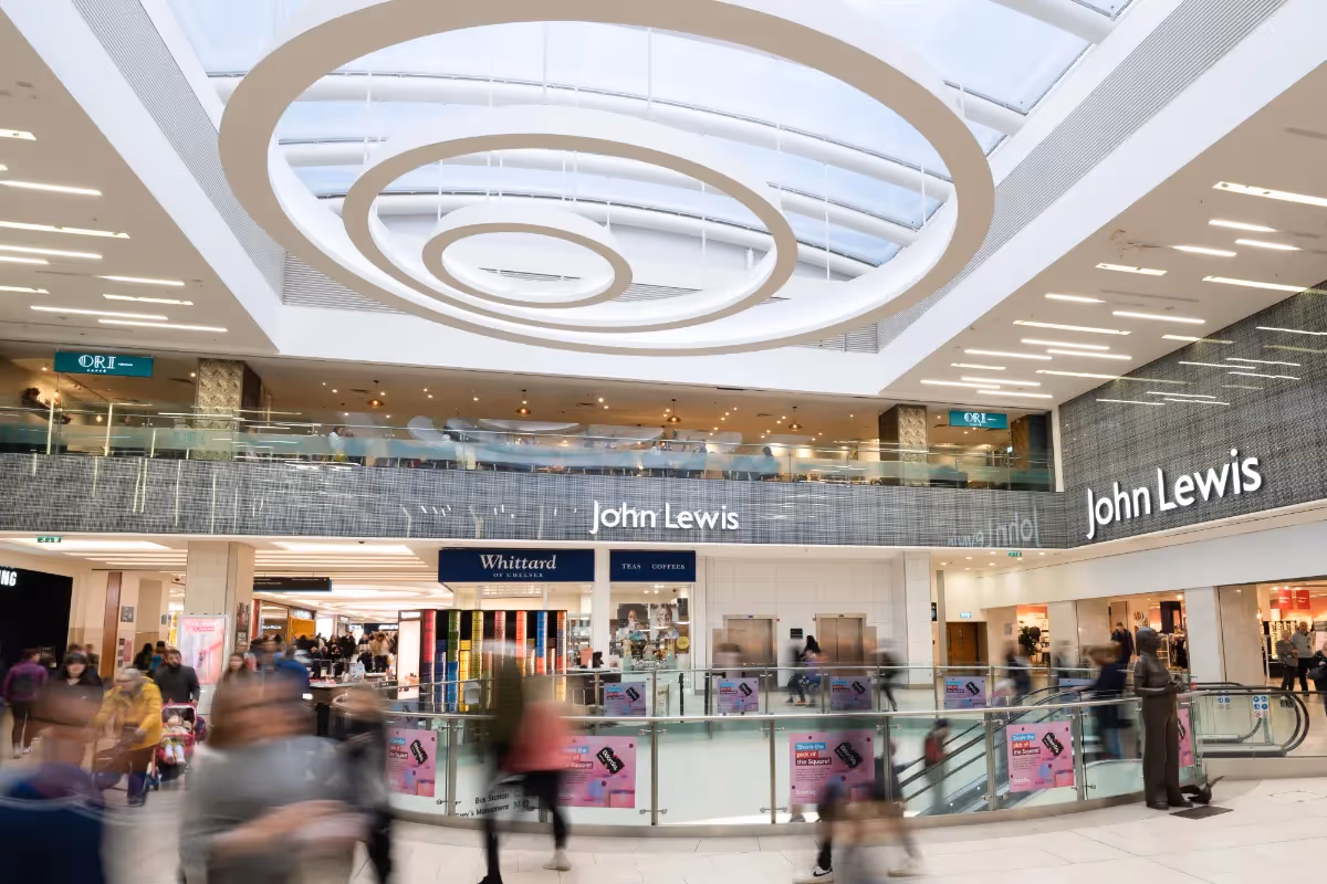 Photographer’s view of a bustling shopping centre: John Lewis shopfront, stylish circular ceiling lights, motion-blurred shoppers passing by.