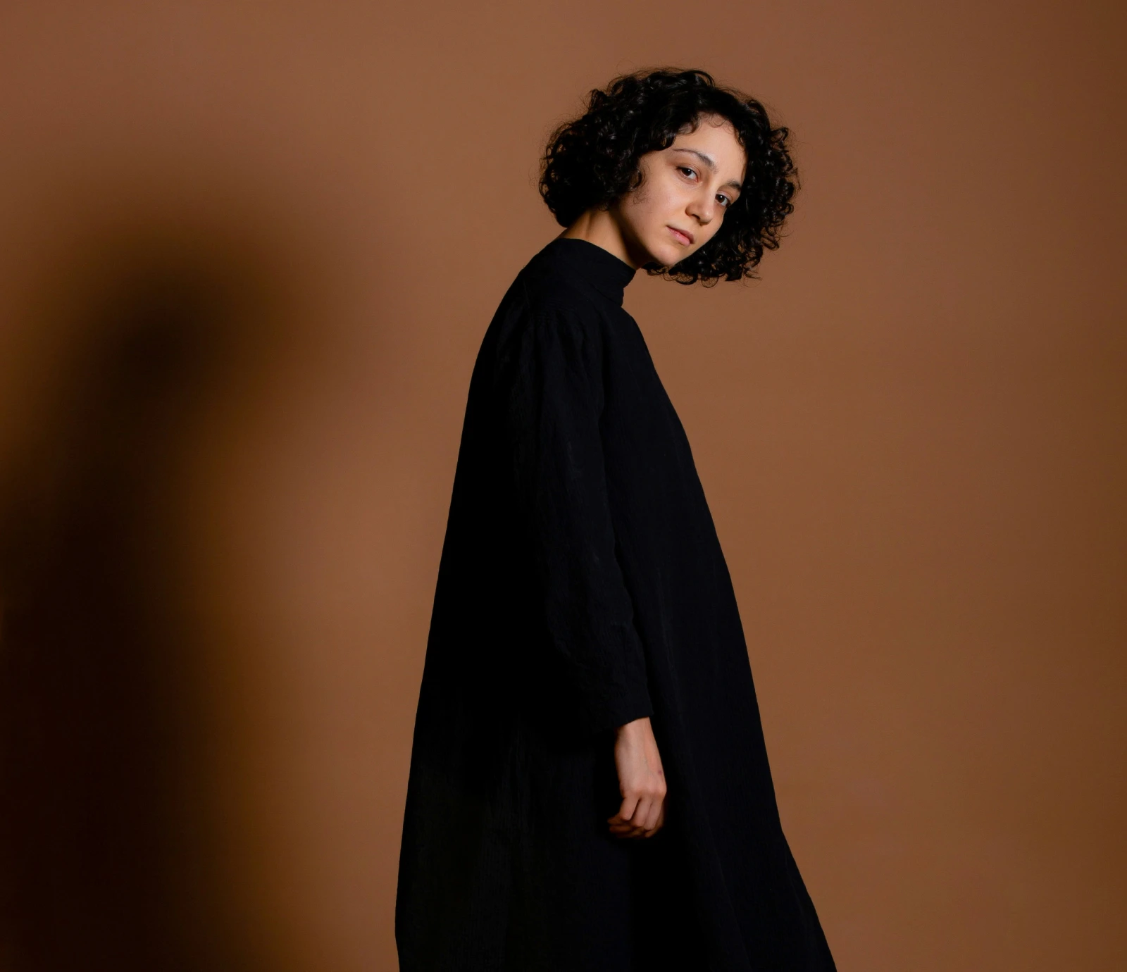 Woman with curly hair wearing a long black dress standing against a brown background.