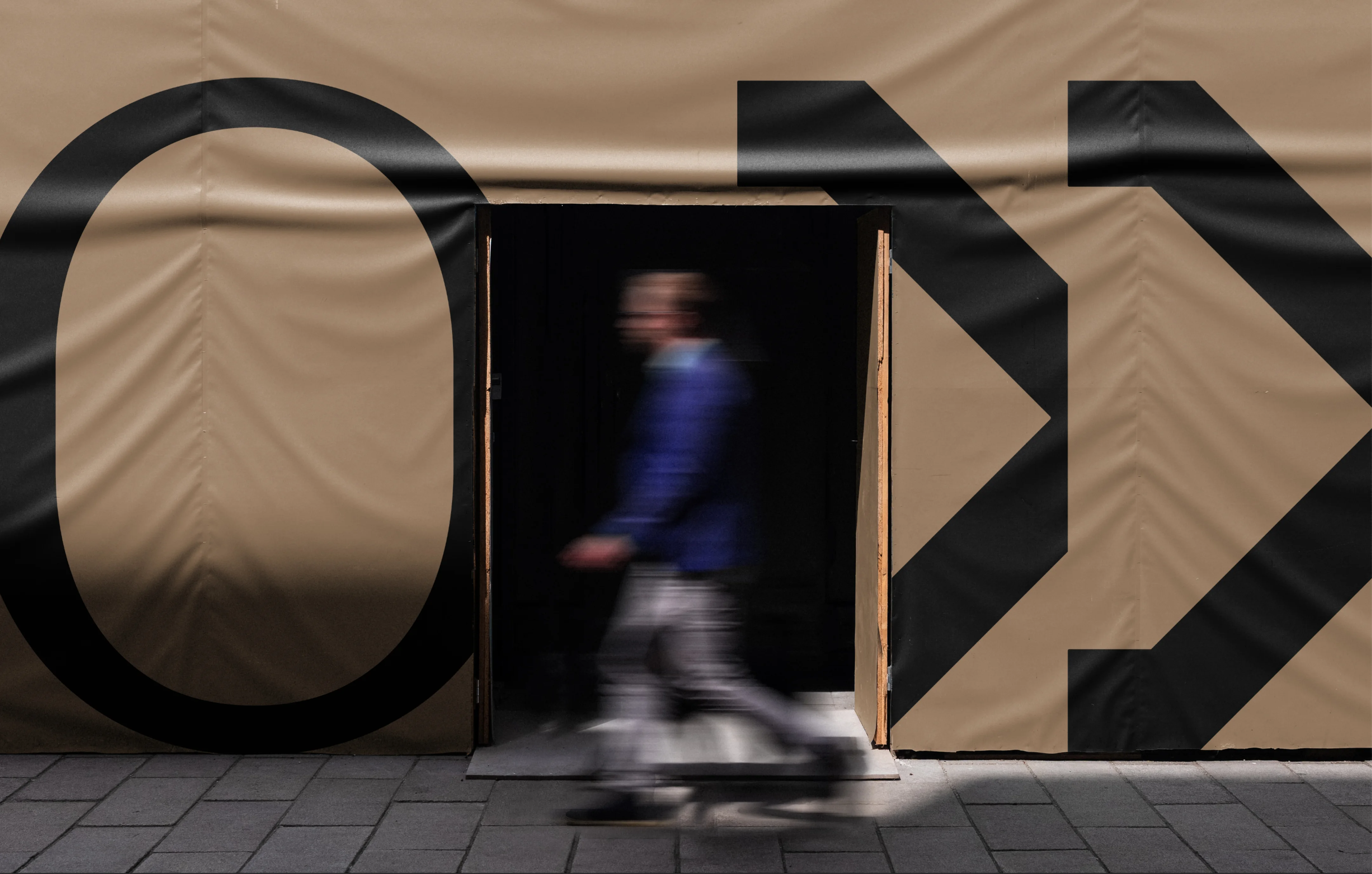 Blurred person wearing a blue jacket walking past a cutout entrance in a beige construction wall with large black letters spelling 'ON'.