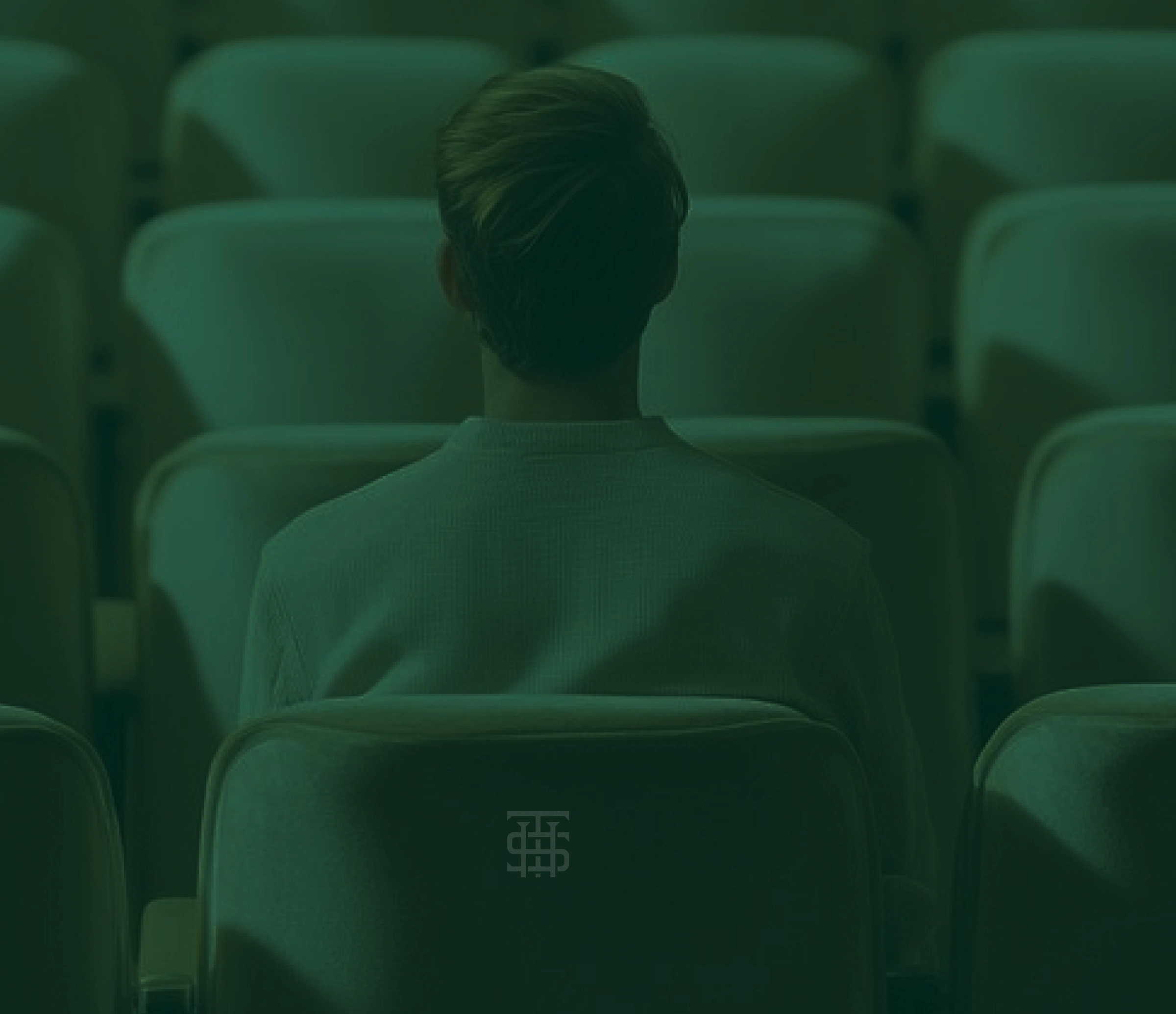 Person wearing a sweater sitting alone facing forward in an empty auditorium with cushioned seats.