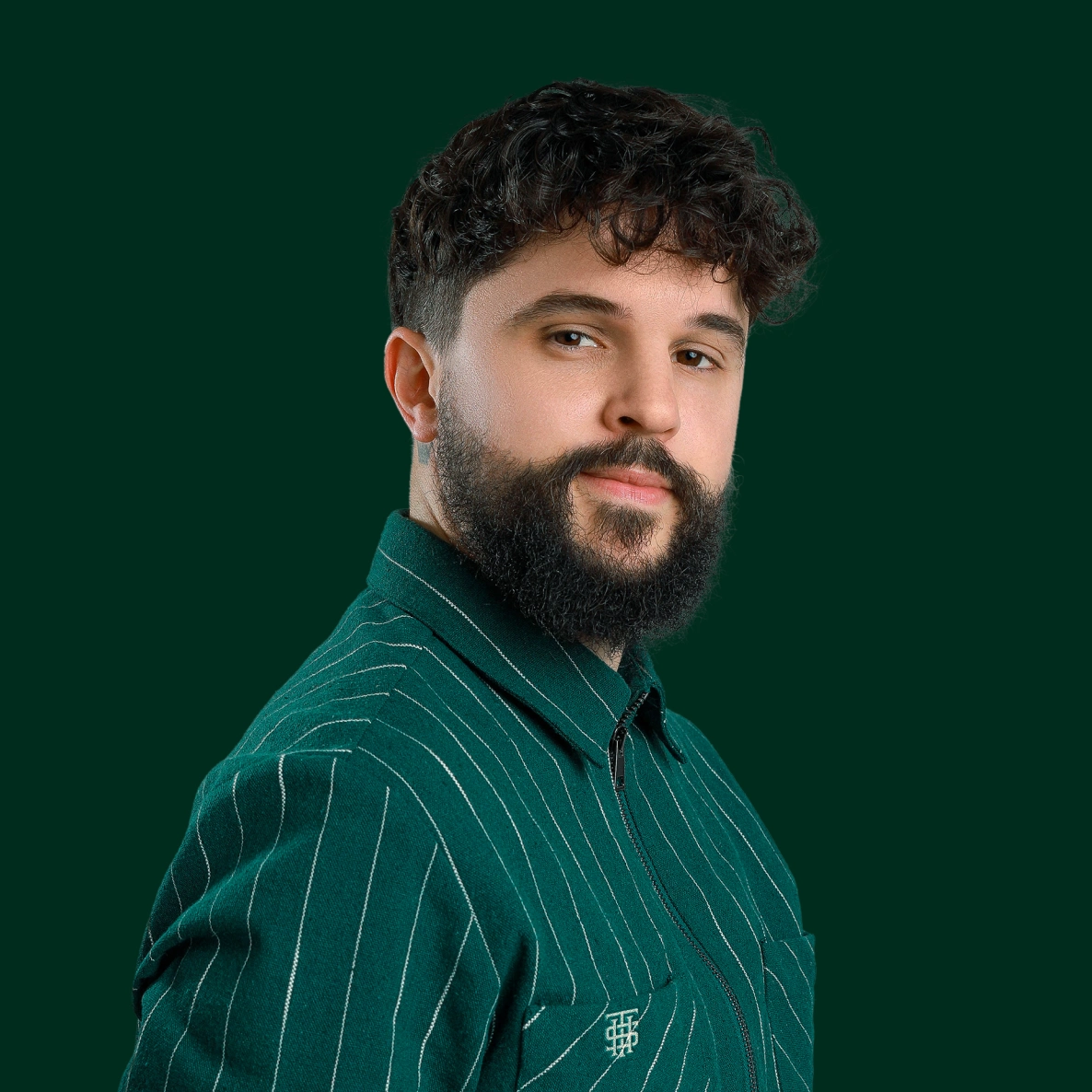 Portrait of a man with curly hair and a full beard wearing a green pinstripe jacket against a dark green background.
