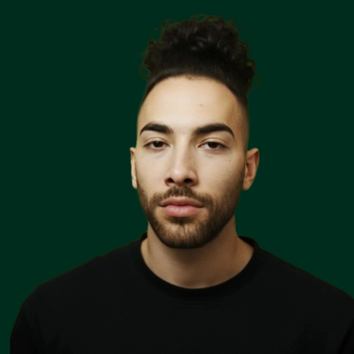 Young man with curly hair tied up, trimmed beard, and wearing a black shirt, against a dark green background.