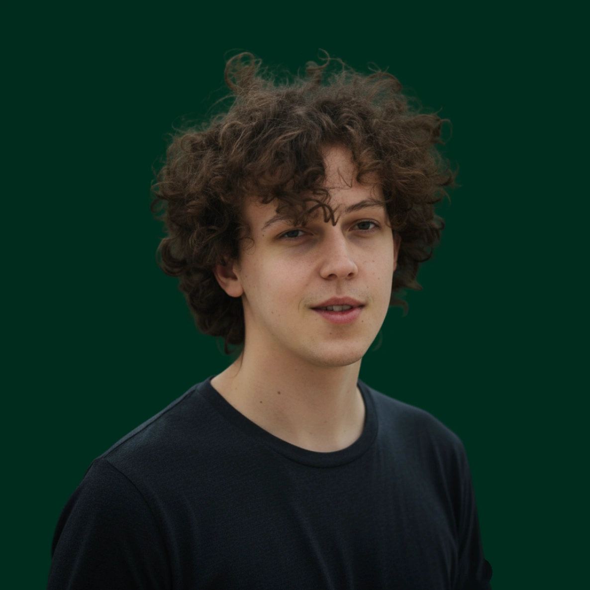 Young man with curly brown hair wearing a black shirt against a dark green background.