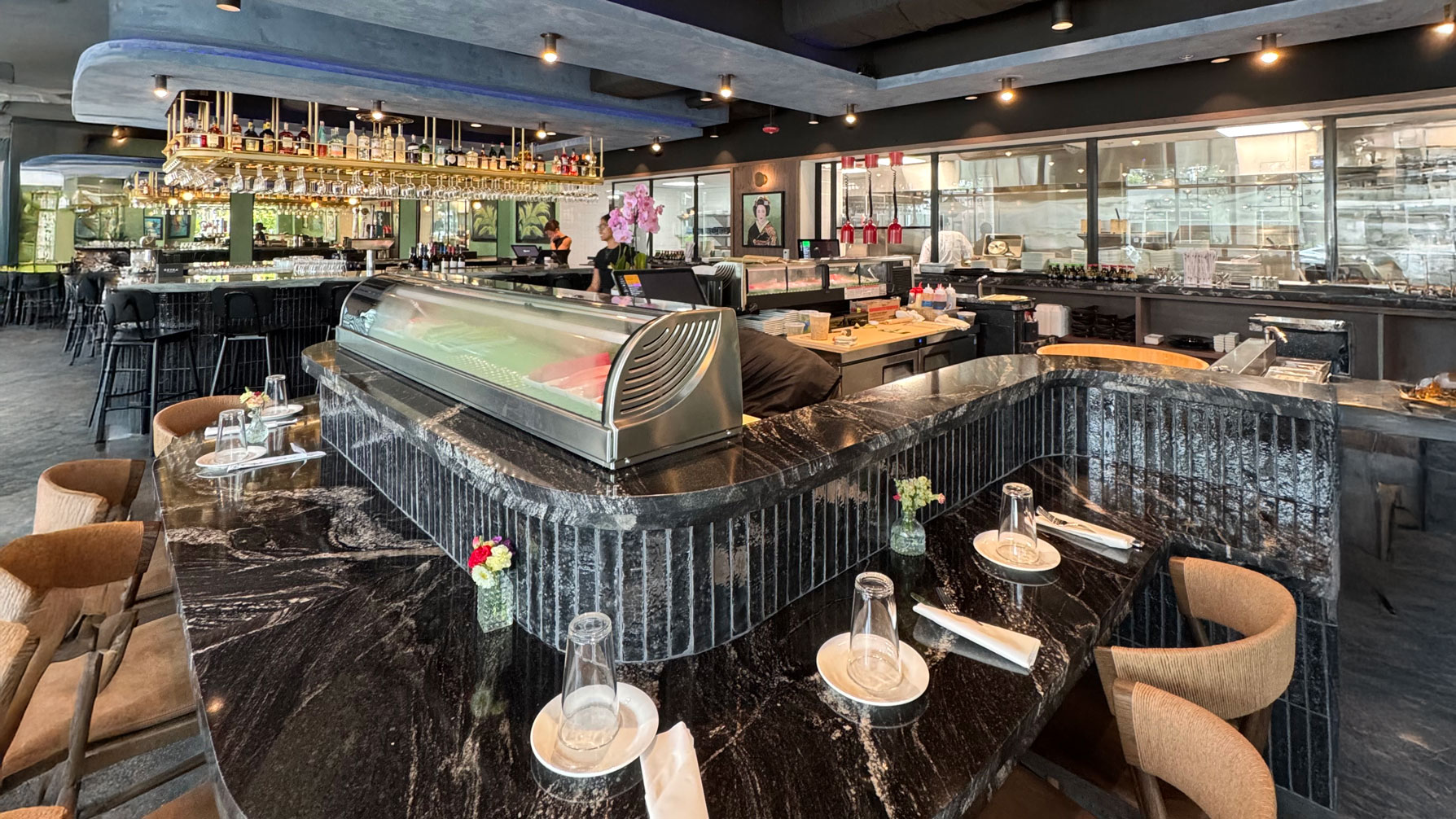 Sleek restaurant interior with a glossy black marble sushi bar surrounded by wooden chairs. Elegant lighting, floral decor, and a vibrant bar area in the background.