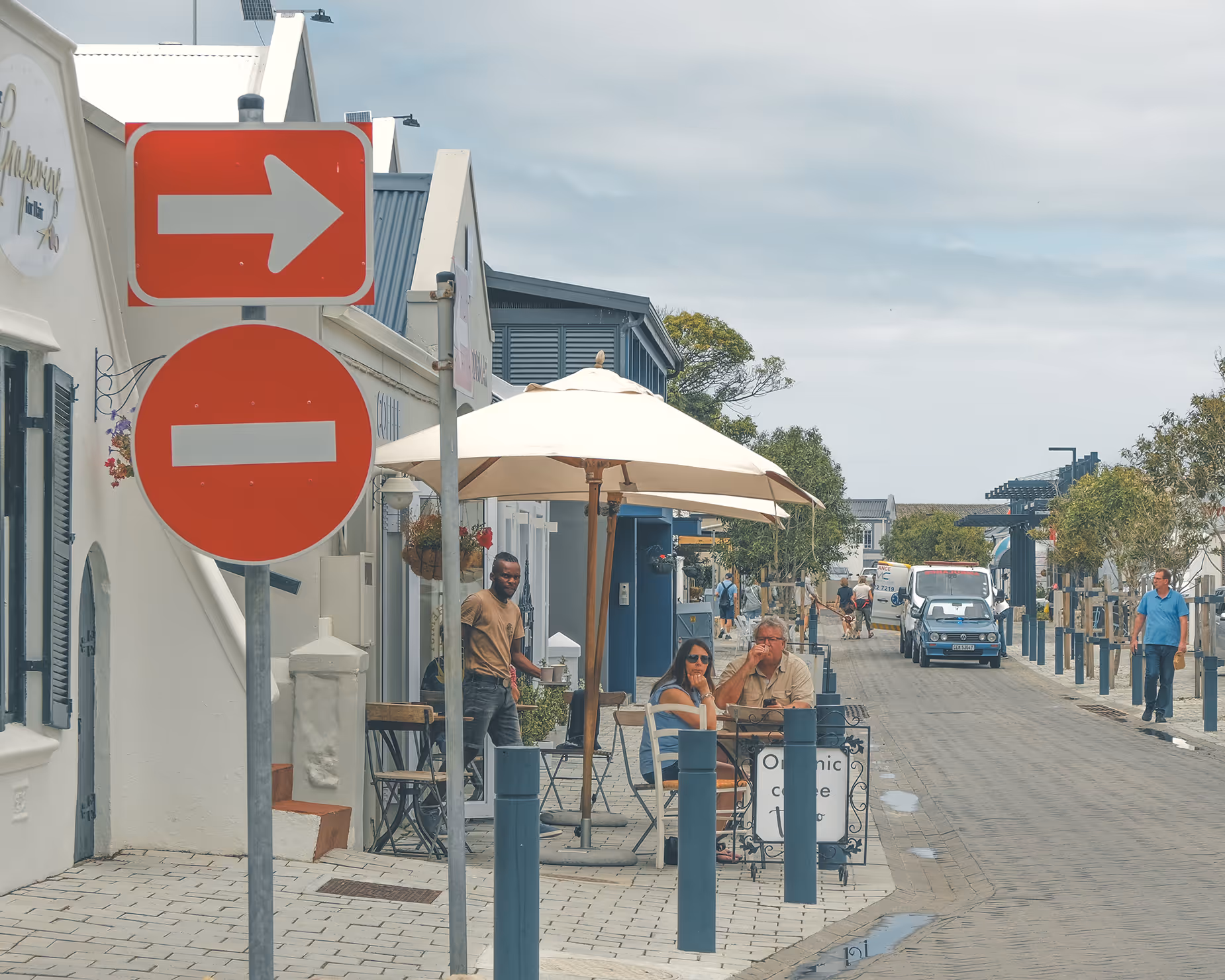 Street view in Hermanus with outdoor seating under umbrellas, people dining and walking, and traffic signs showing right turn and no entry.