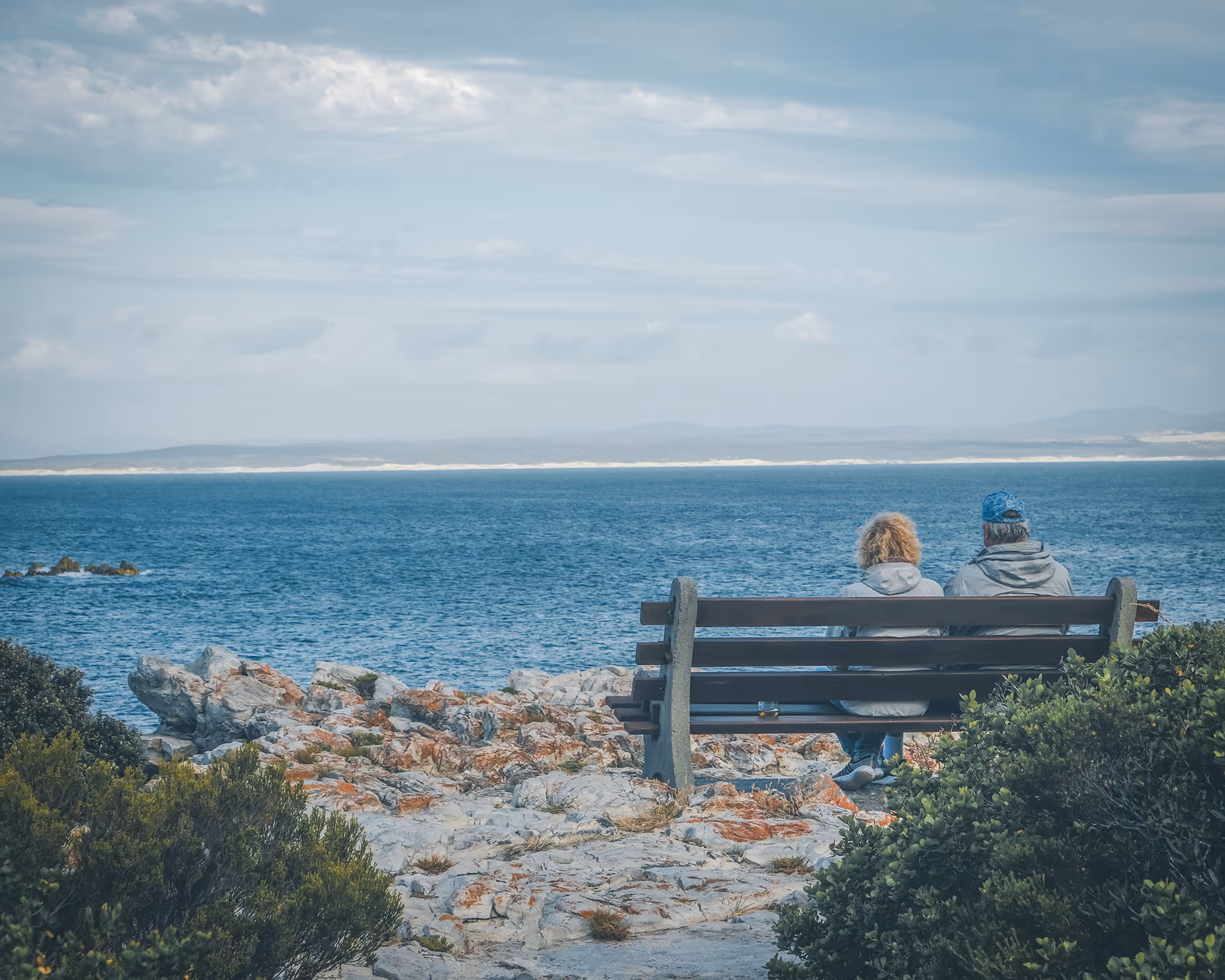 Two people sitting on a bench overlooking a rocky shore and a blue ocean under a cloudy sky.