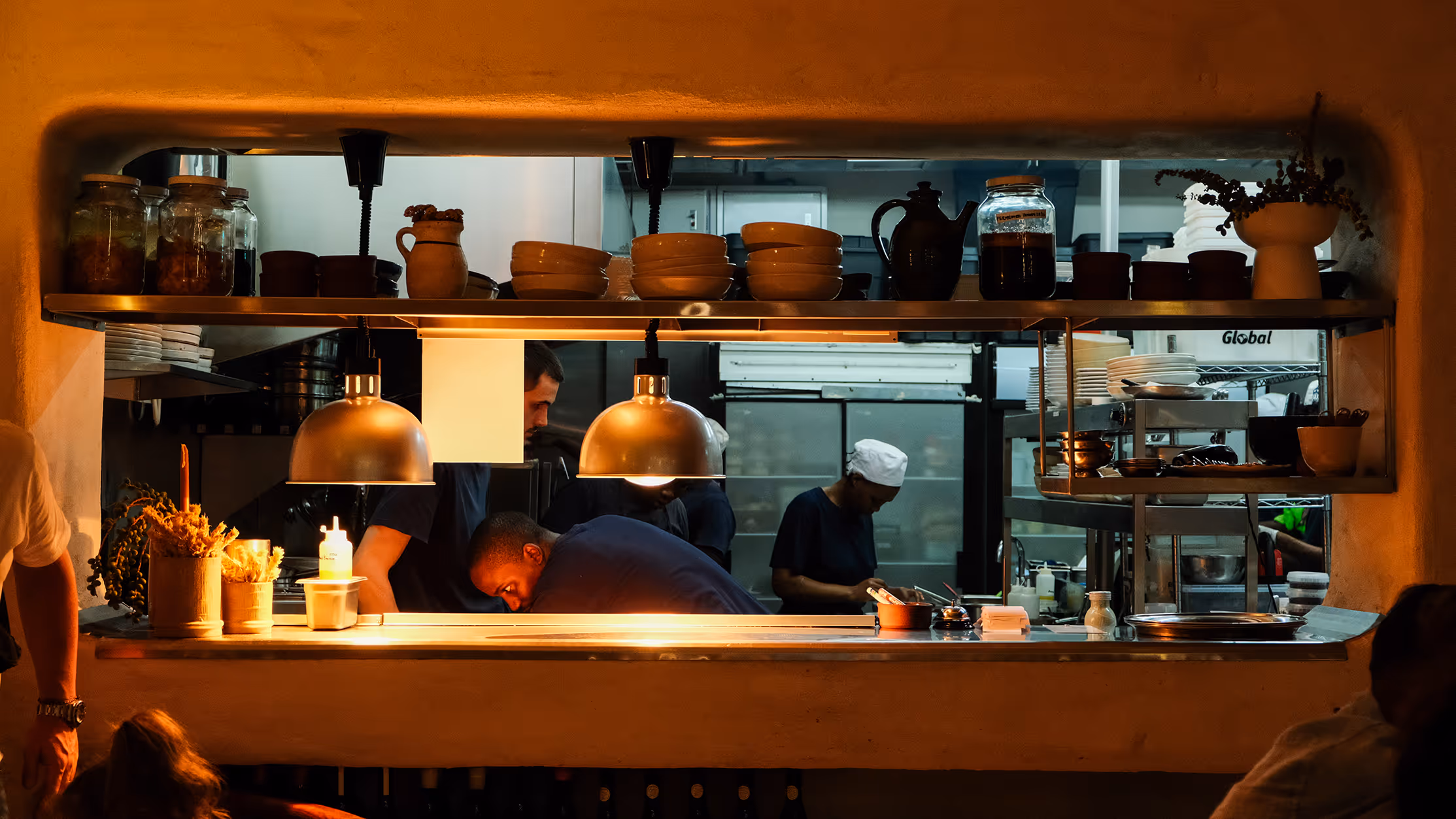Busy restaurant kitchen pass with chefs working under warm hanging lamps and shelves holding bowls, jars, and plants.