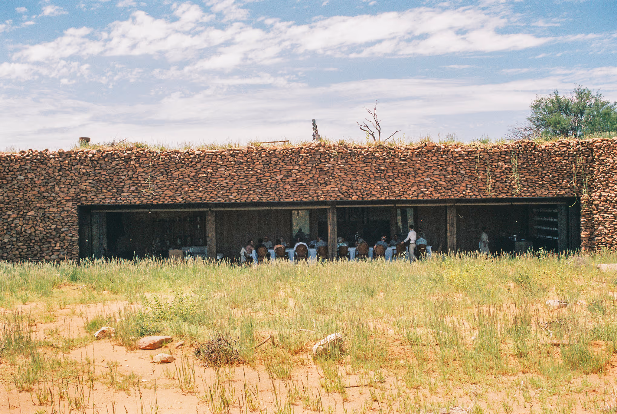 Stone building with people dining inside, seen through a wide open facade, surrounded by grass and under a partly cloudy sky.