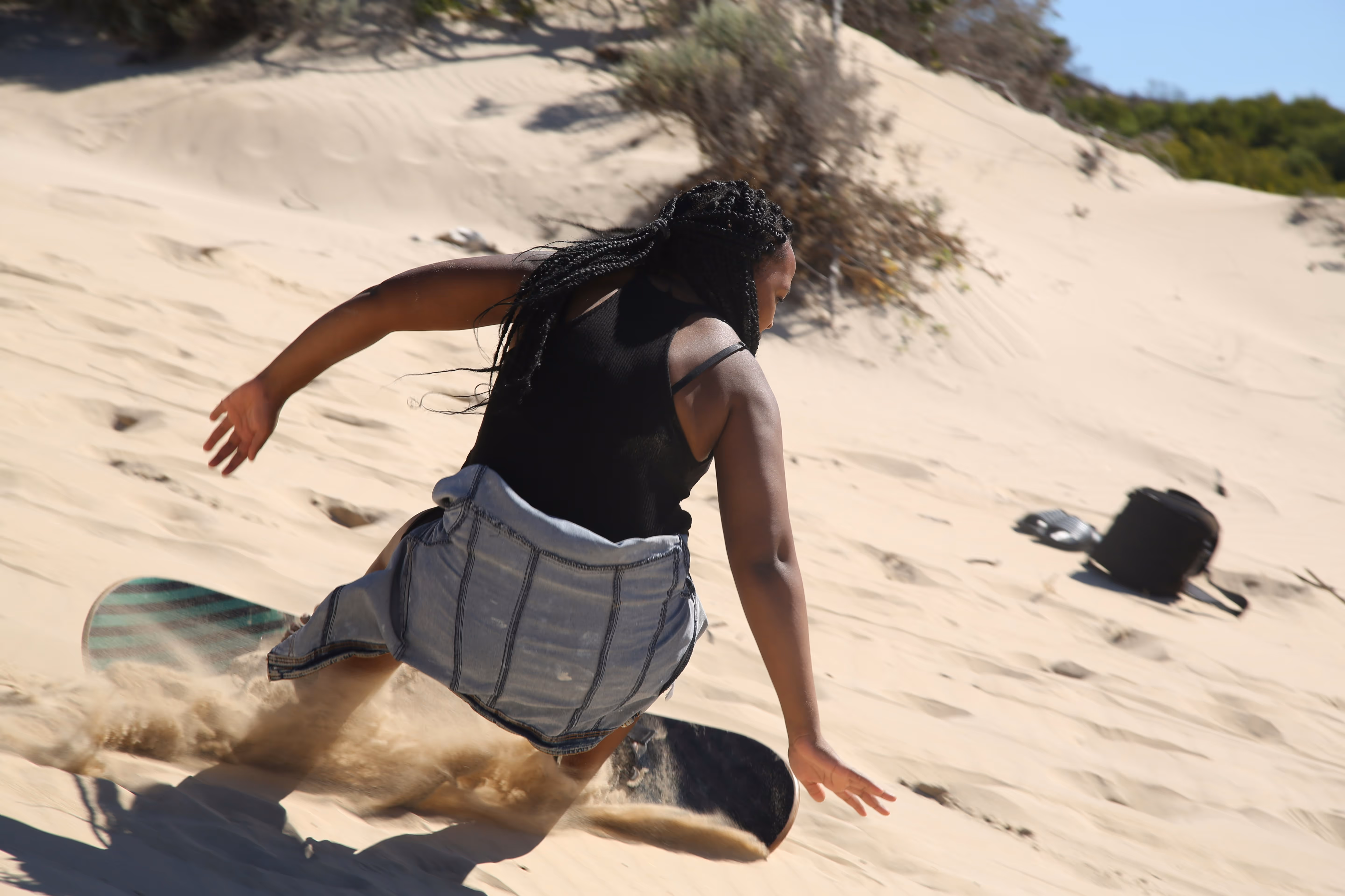 Person with braided hair wearing a black top and denim skirt sandboarding down a sandy dune.