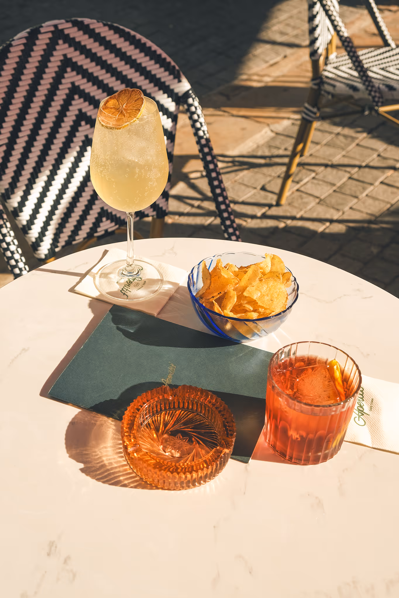Outdoor table with two cocktails, a bowl of potato chips, and an orange glass ashtray in sunlight.