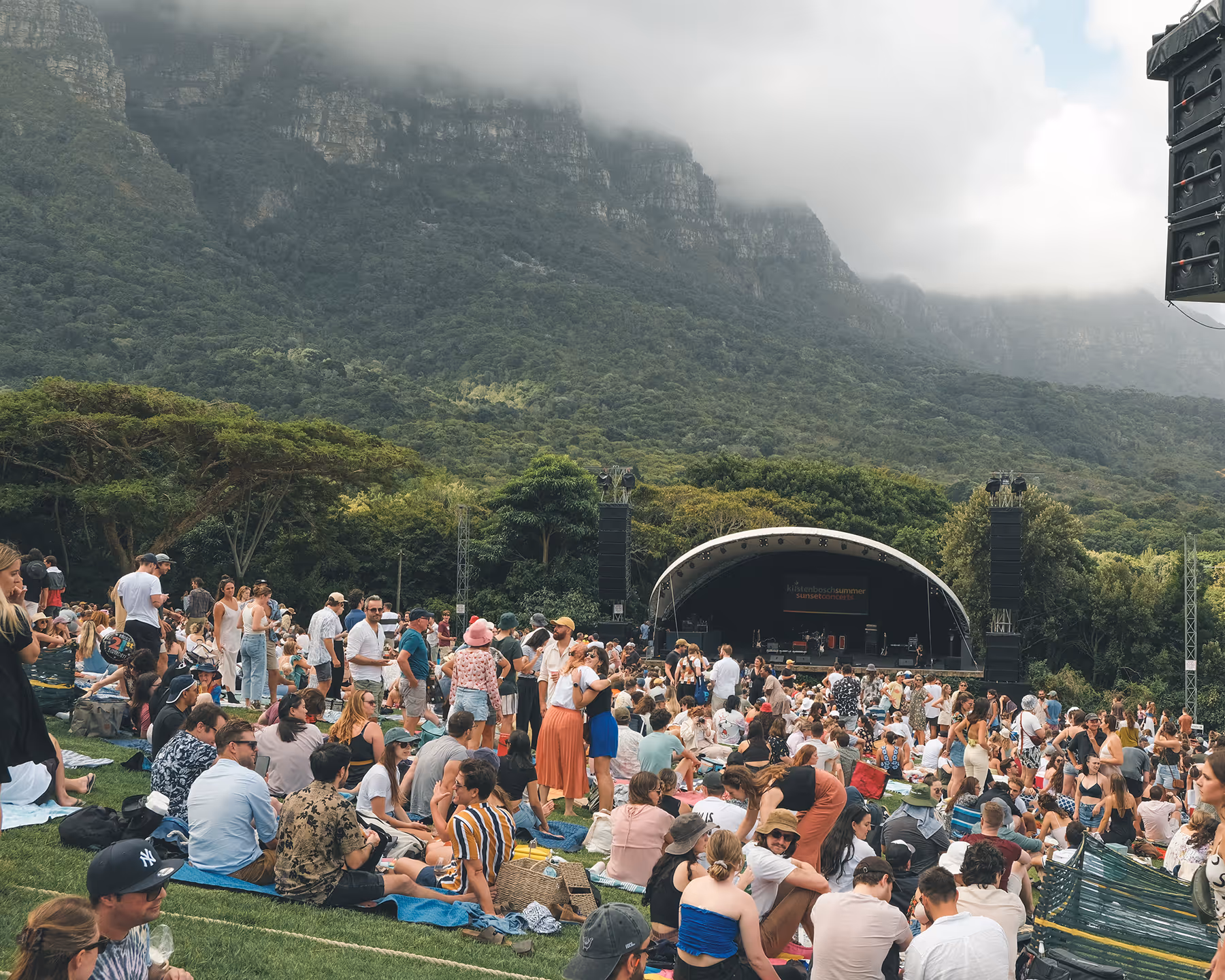 Crowd gathered on a grassy hillside with a stage set up for an outdoor concert at Kirstenbosch Gardens, with misty mountains in the background.