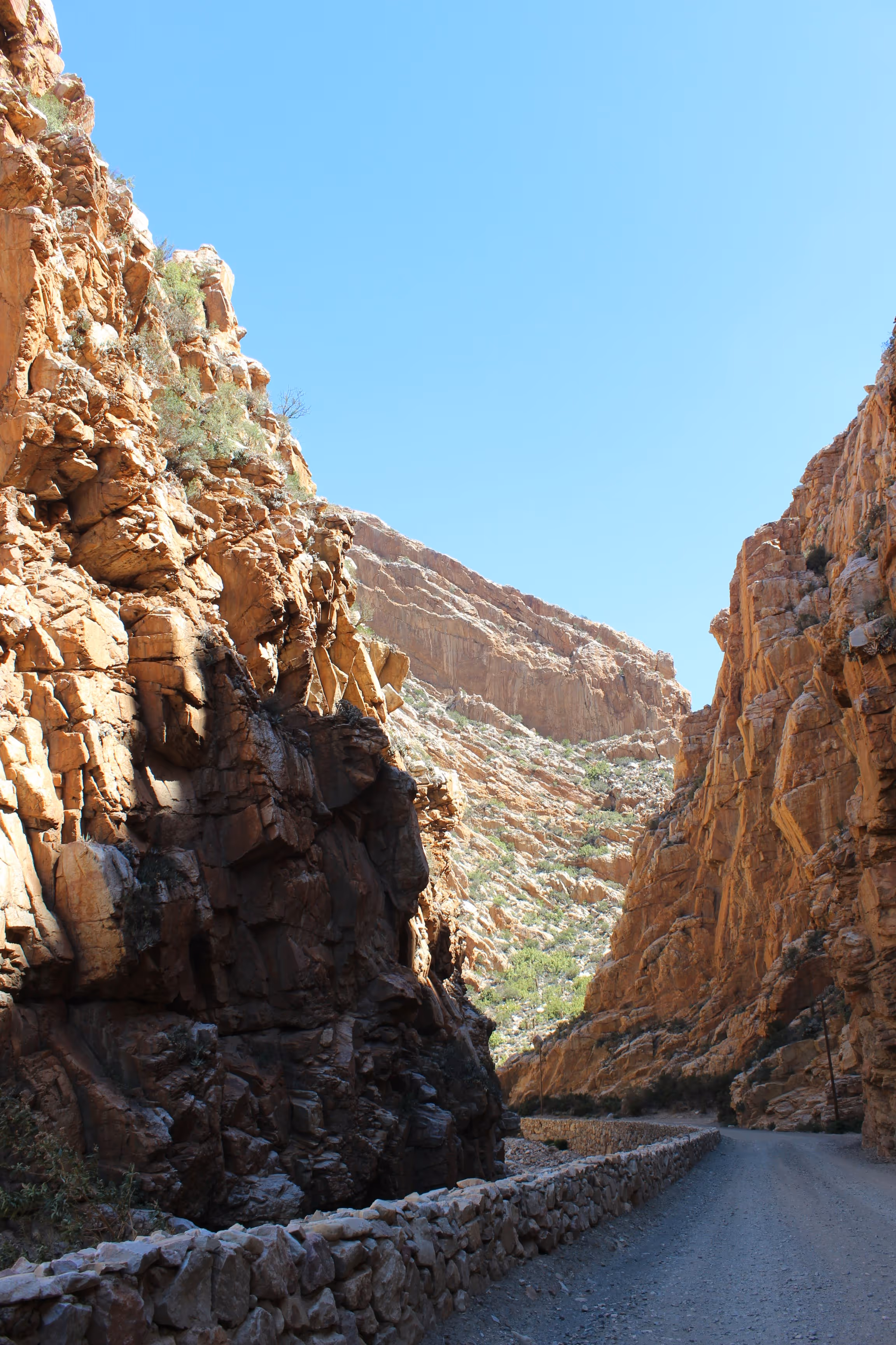 Winding gravel road flanked by tall rocky cliffs under a clear blue sky.