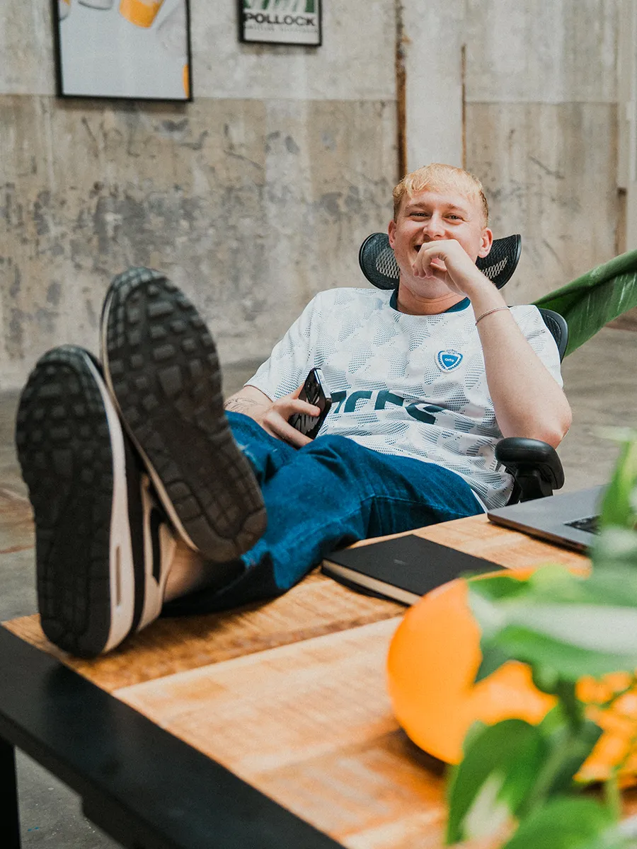 Jonge man zit lachend achter een bureau in een industriële studio, met zijn voeten op tafel en een smartphone in de hand. Op de achtergrond zijn betonnen muren en een kunstposter zichtbaar.