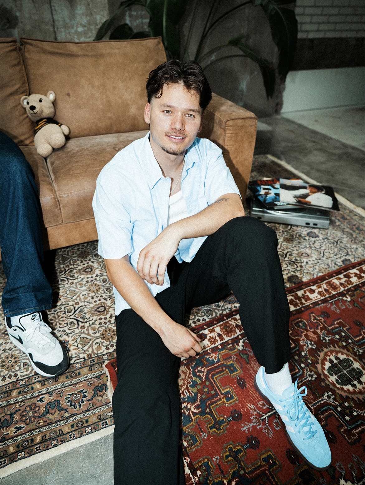 Jonge man zit op een Perzisch vloerkleed in een creatieve studio, met een bruine bank, een teddybeer en een platenspeler met platen op de achtergrond.