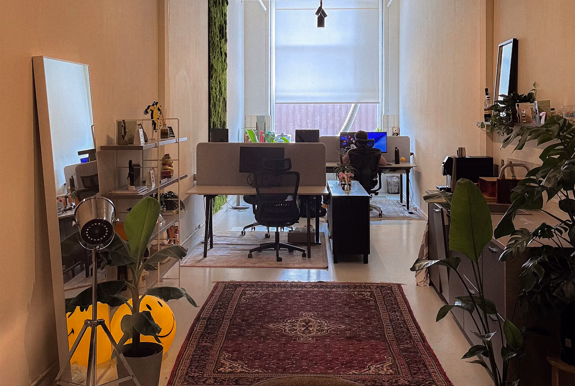 Kantoorruimte met bureau en computerschermen bij een raam, een persoon zittend voor een computer, planten en een grote spiegel met gele smileyballonnen op de vloer, en een rood tapijt op de vloer.