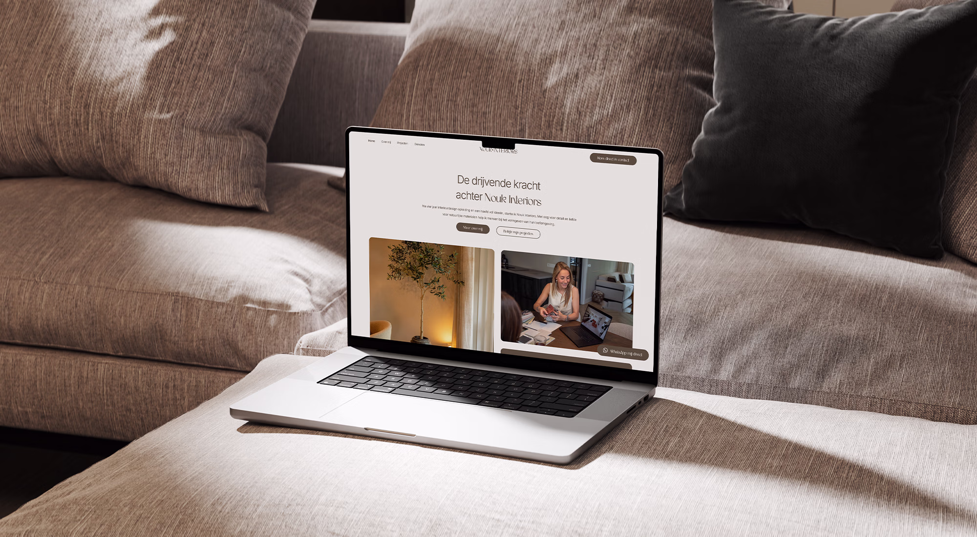 Laptop met een interieurwebsite geopend, geplaatst op een beige stoffen bank met kussens, onder natuurlijk licht.