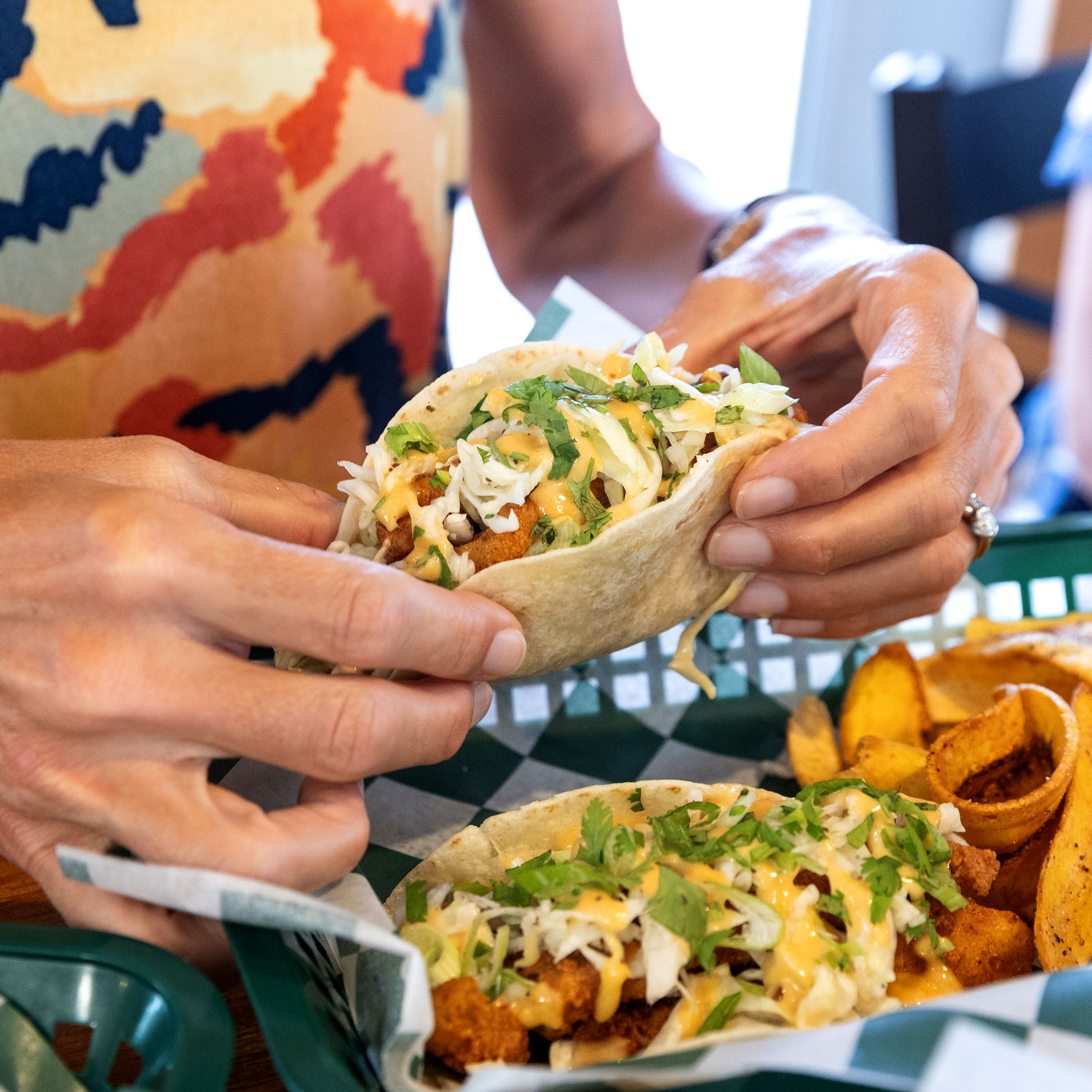 Hands holding a fish taco topped with shredded cabbage, cilantro, and creamy sauce, with a basket of tacos and seasoned potato wedges on a checkered paper-lined tray.