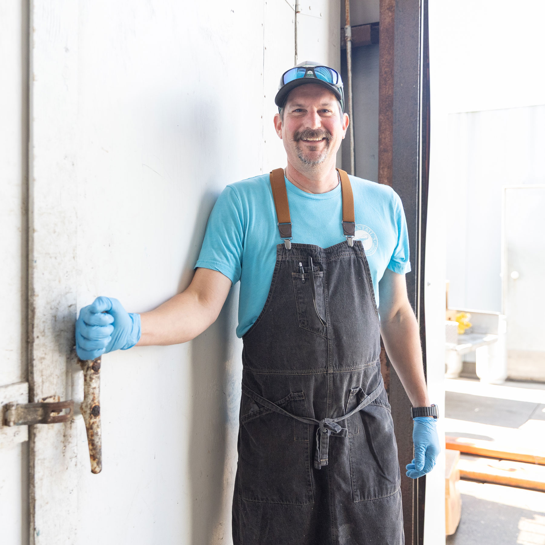 Man wearing blue gloves, a blue shirt, a dark apron, and a cap smiling while holding open a door in a bright industrial setting.