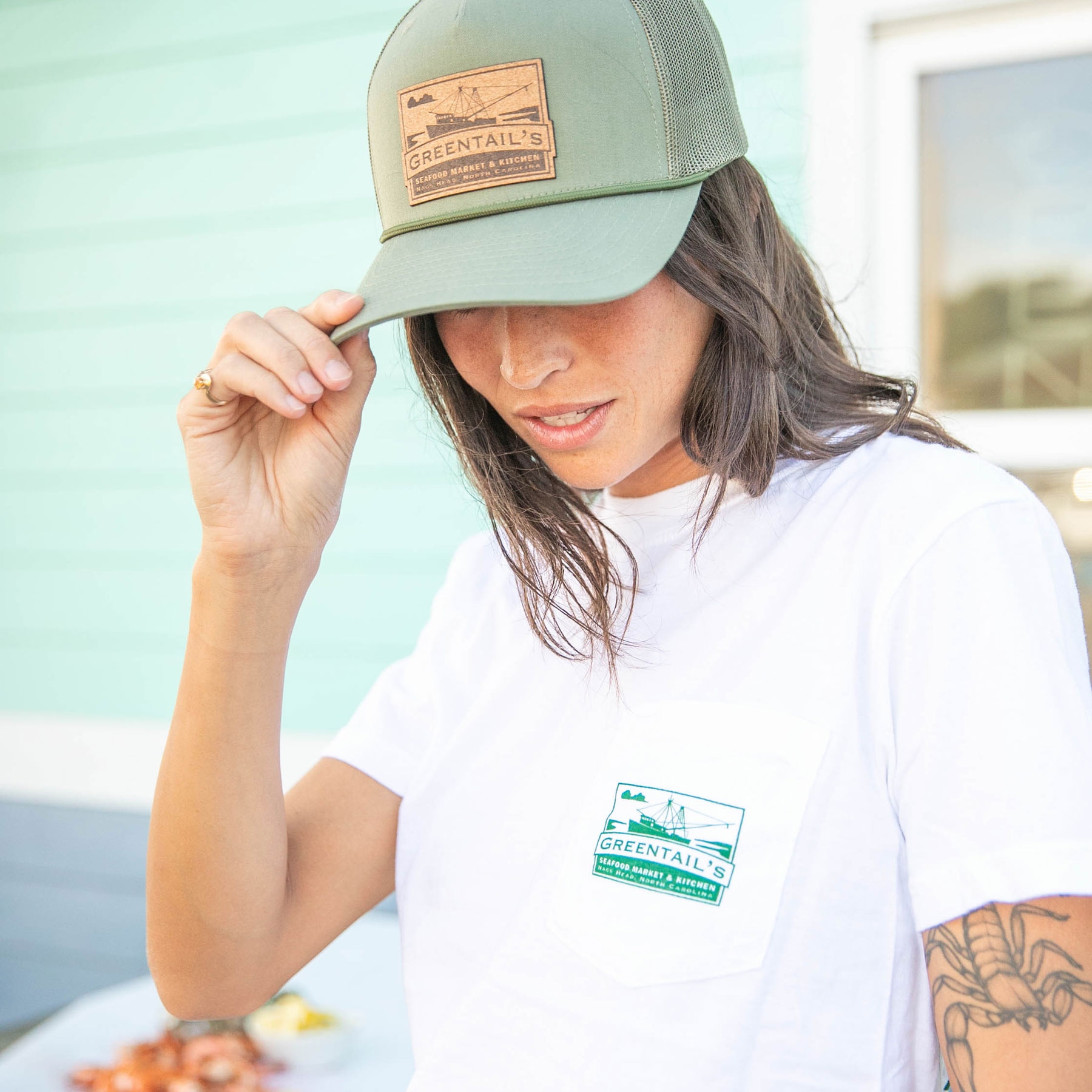Woman wearing a green GREENTAIL'S cap and white t-shirt with a GREENTAIL'S logo on the pocket, adjusting the cap brim.