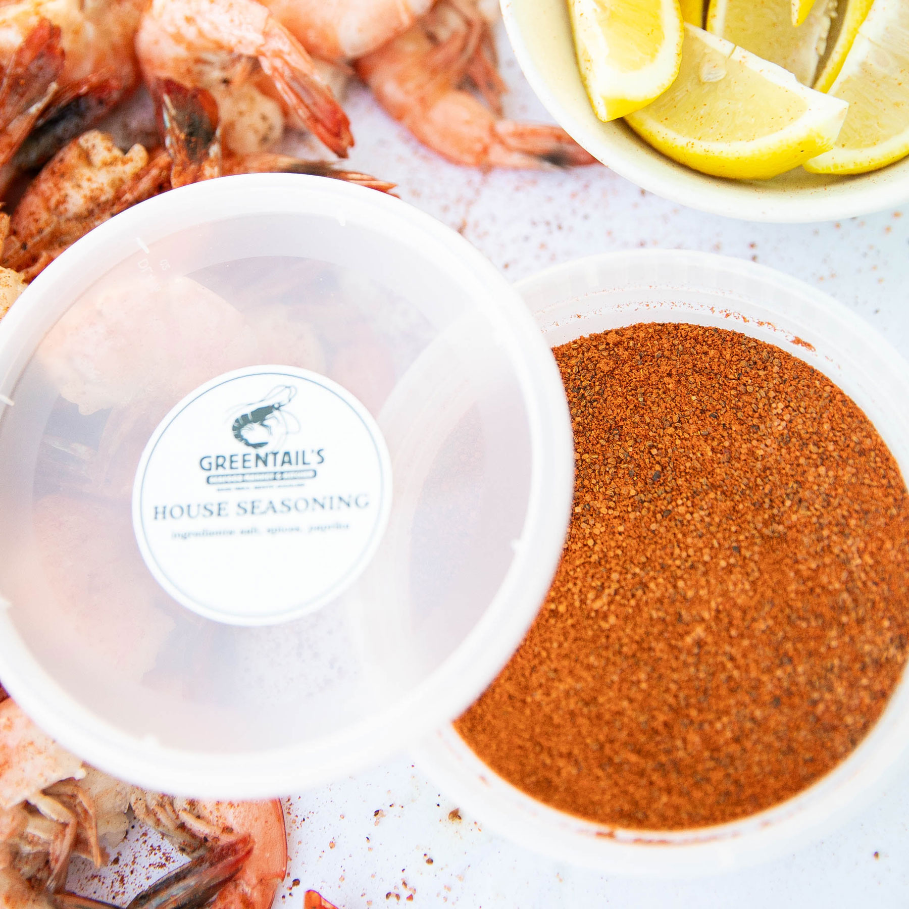 Container of Greentail's house seasoning spice blend next to cooked shrimp and a bowl of lemon wedges.