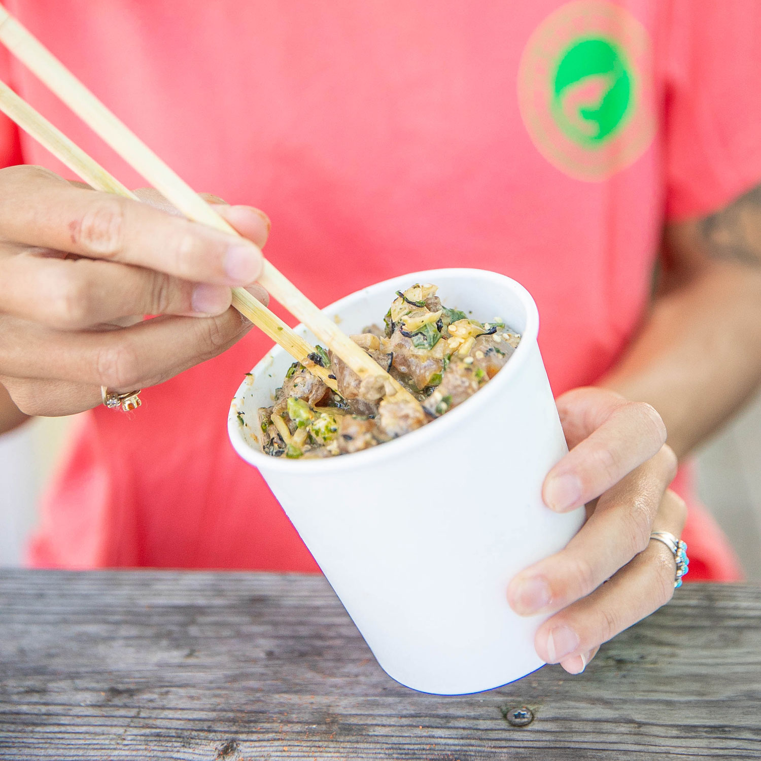 Person wearing a red shirt using chopsticks to eat a mixed dish from a white paper cup.