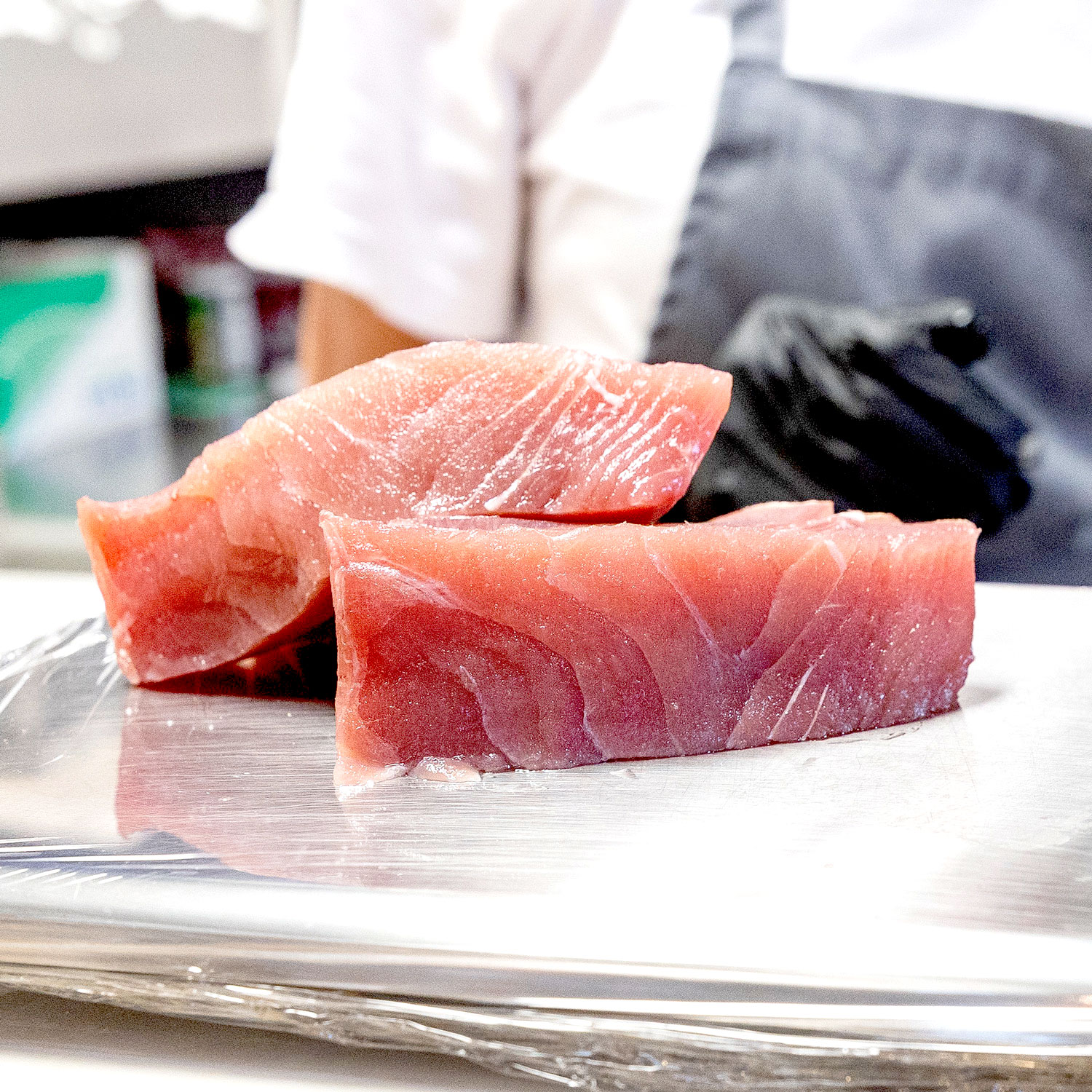 Two pieces of raw tuna sashimi on a clear plastic-wrapped surface with a person wearing a white shirt and black glove in the background.