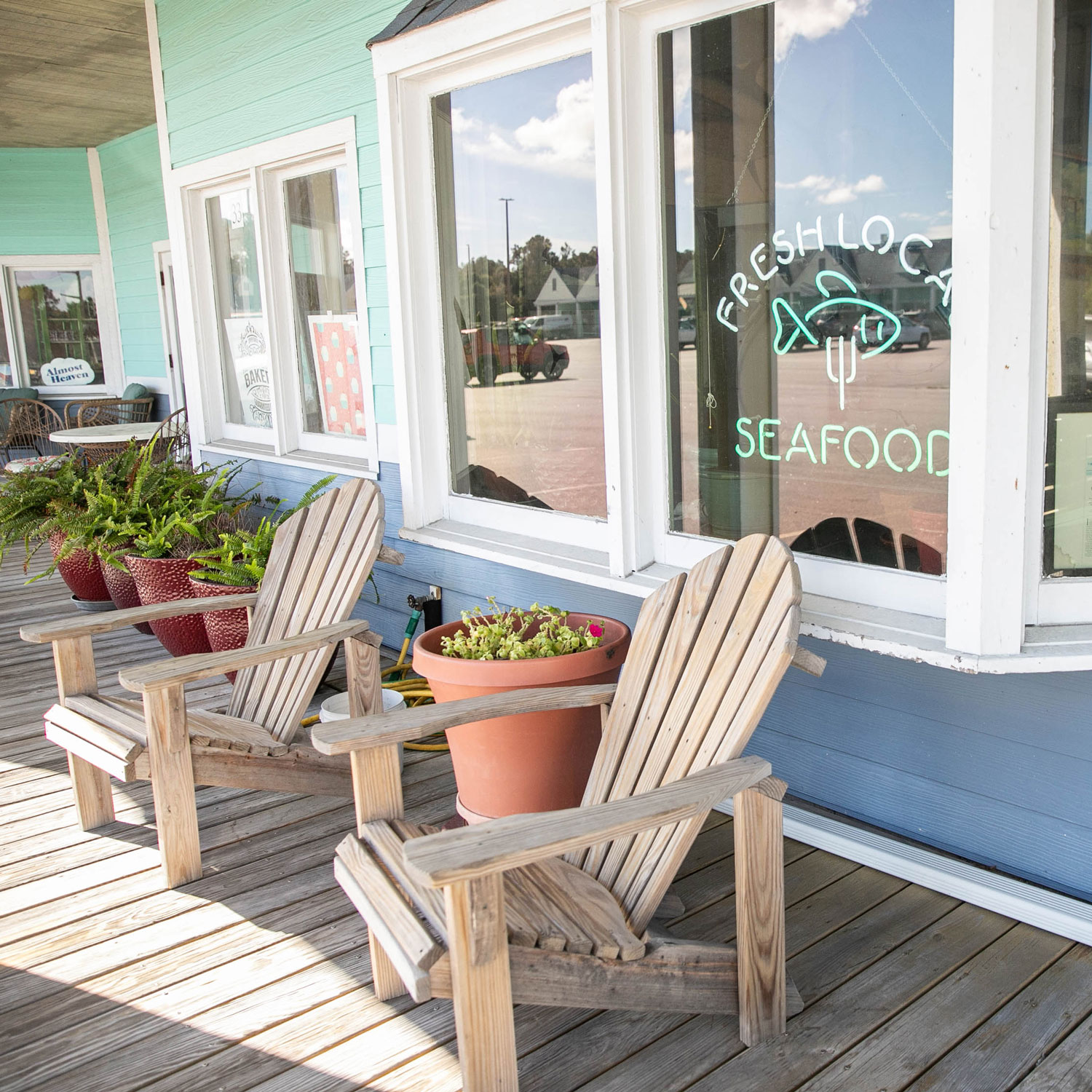 Wooden Adirondack chairs and potted plants on a porch outside a seafood restaurant with a neon sign.