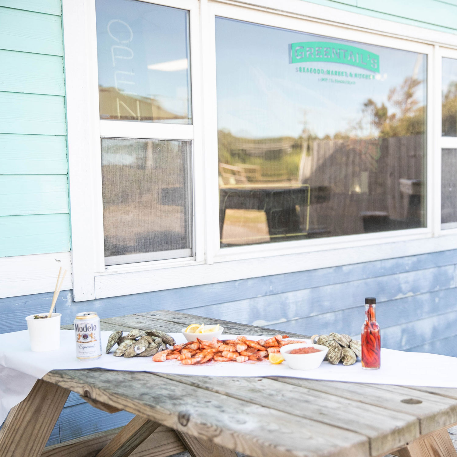 Outdoor wooden picnic table with cooked shrimp, oysters, lemon wedges, cocktail sauce, hot sauce, and a can of Modelo beer in front of a seafood market window.