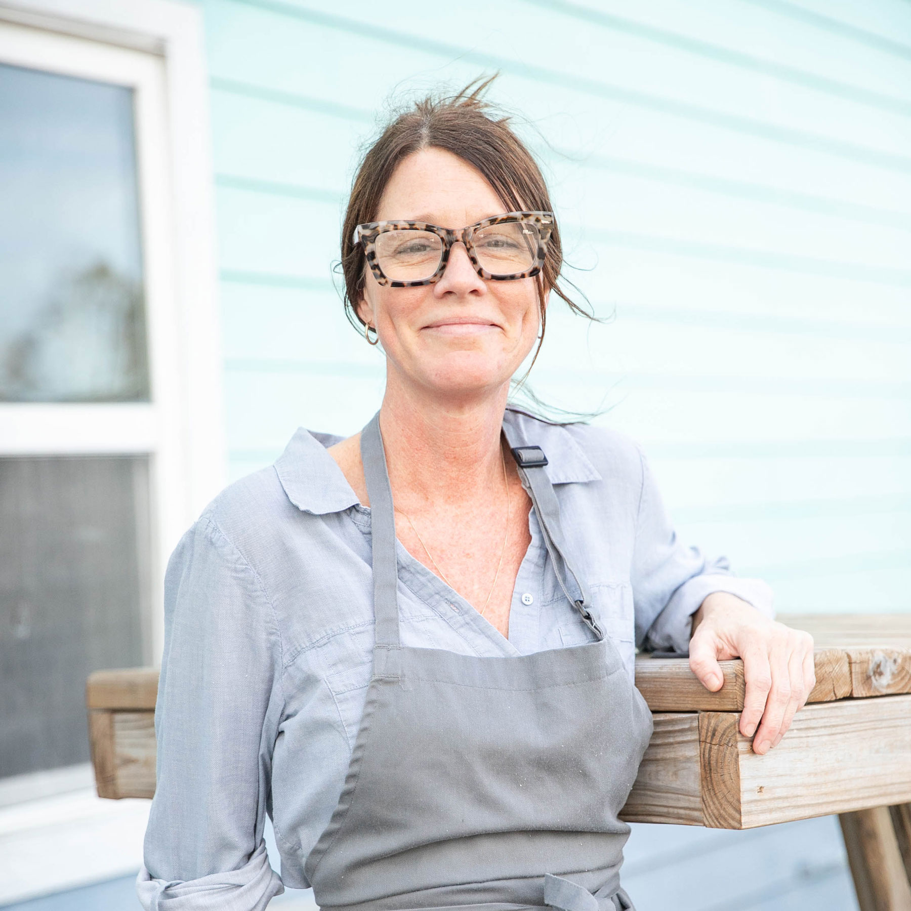 Smiling woman with glasses and gray apron leaning on a wooden table outside a light blue building.