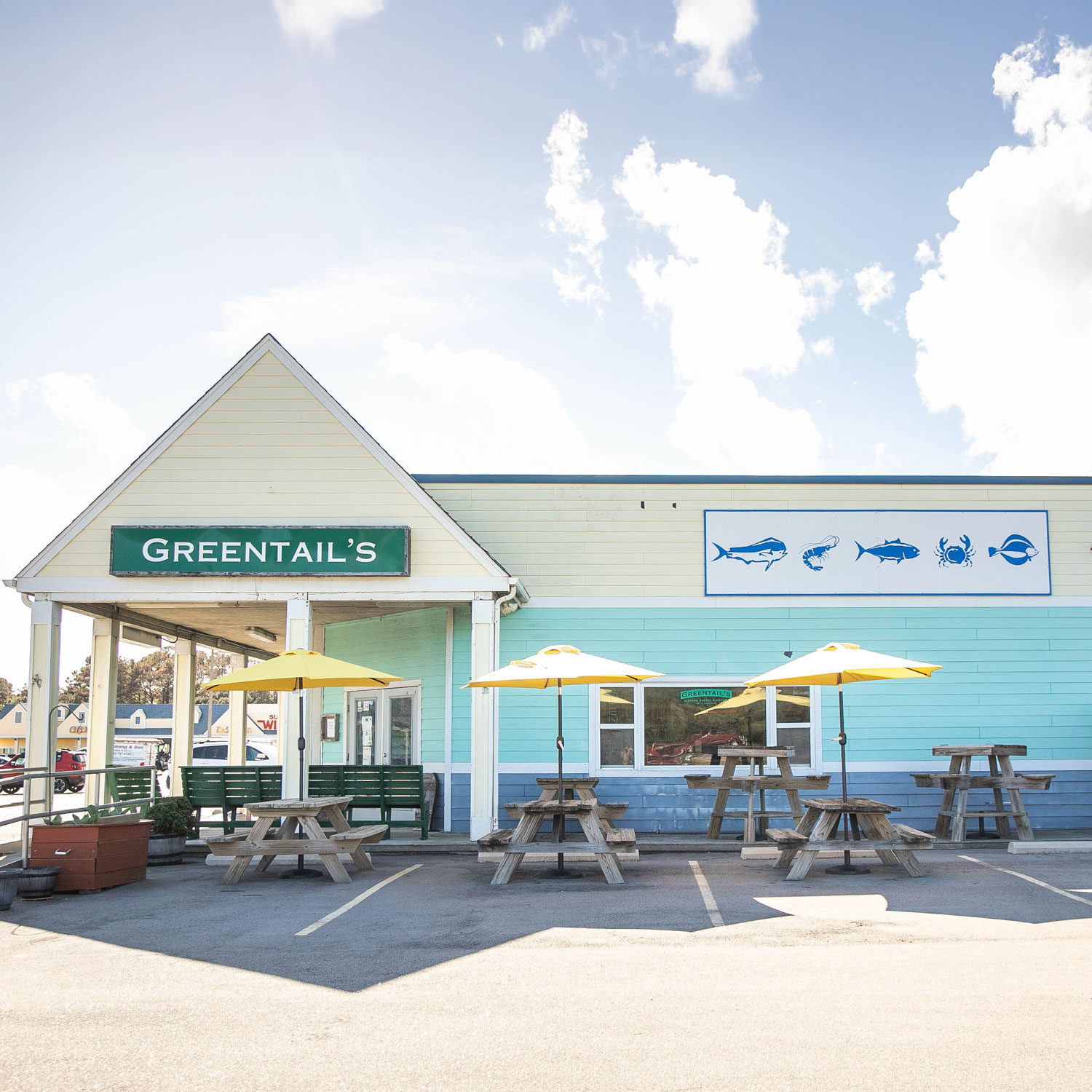 Exterior of Greentail's seafood restaurant with picnic tables and yellow umbrellas in a parking lot under a partly cloudy sky.