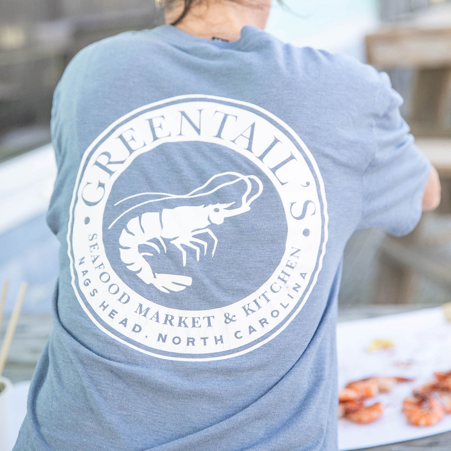 Person wearing a blue shirt with Greentails Seafood Market & Kitchen logo featuring a shrimp and location Nags Head, North Carolina.
